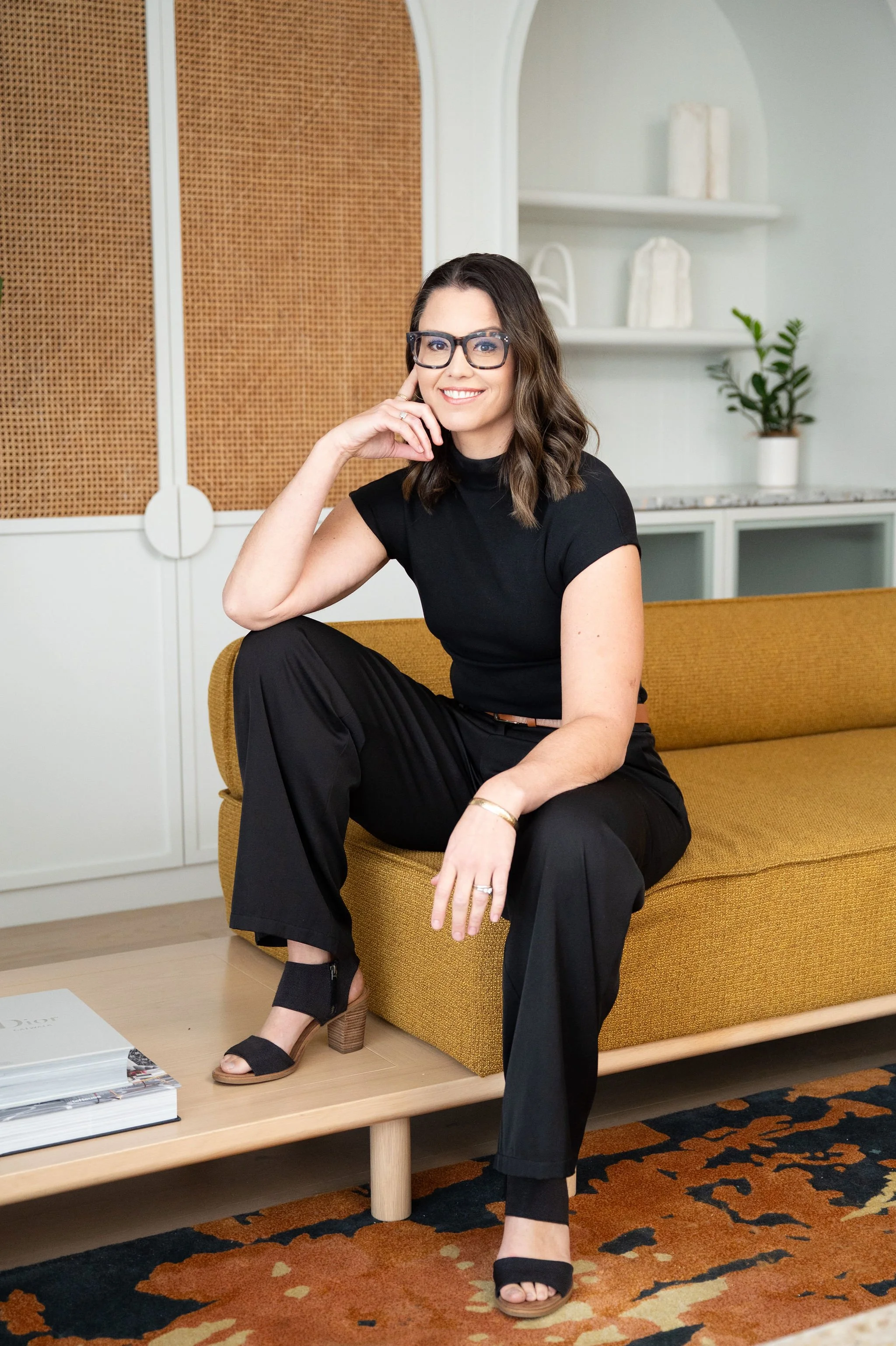 A woman with dark brown hair, glasses, and a black shirt sitting on a mustard yellow sofa in a room with white and brown decor, including books, a potted plant, and patterned carpet.