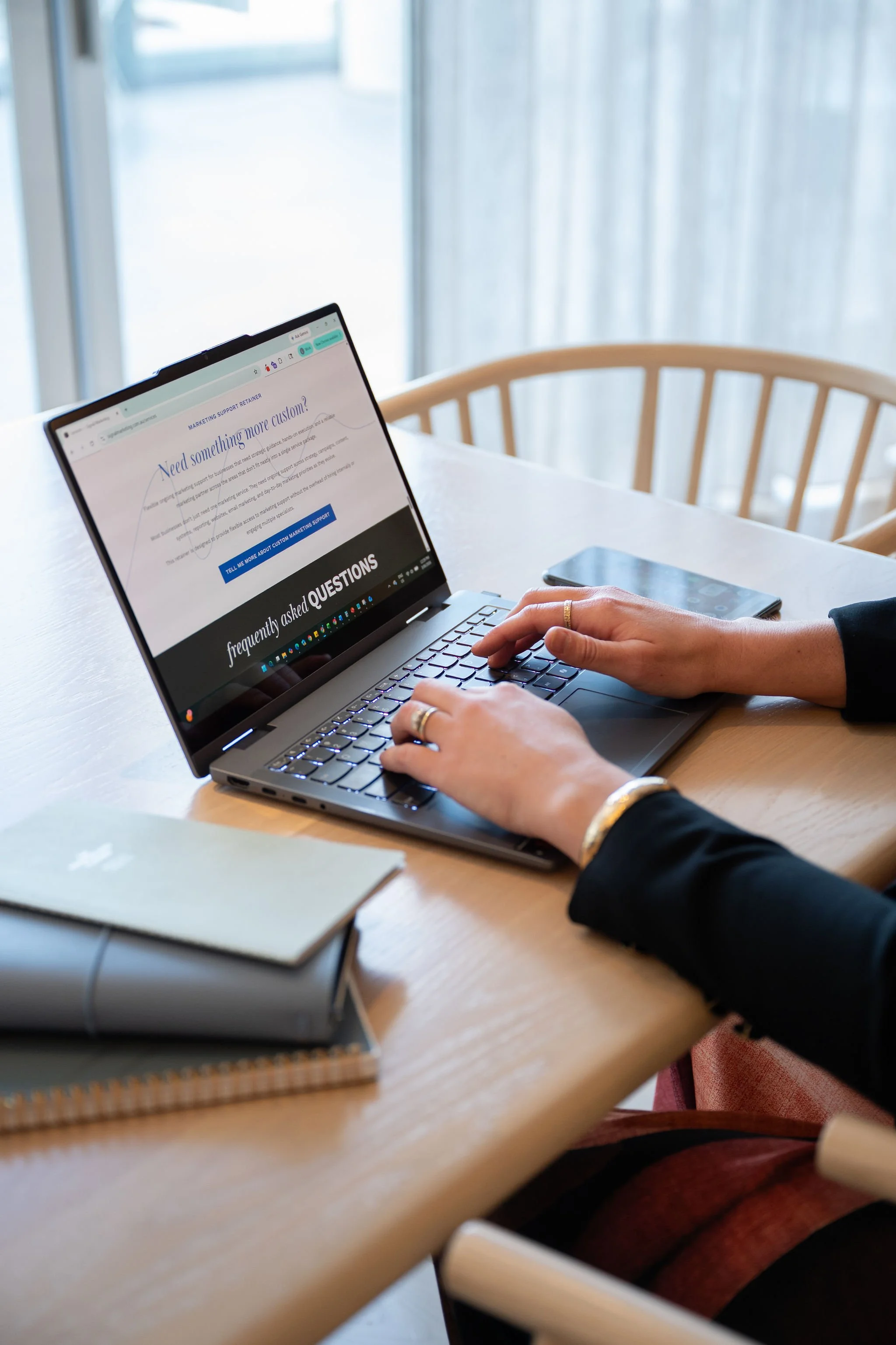 Person typing on a silver laptop with a webpage about marketing support, a smartphone next to it, and notebooks on a wooden table.