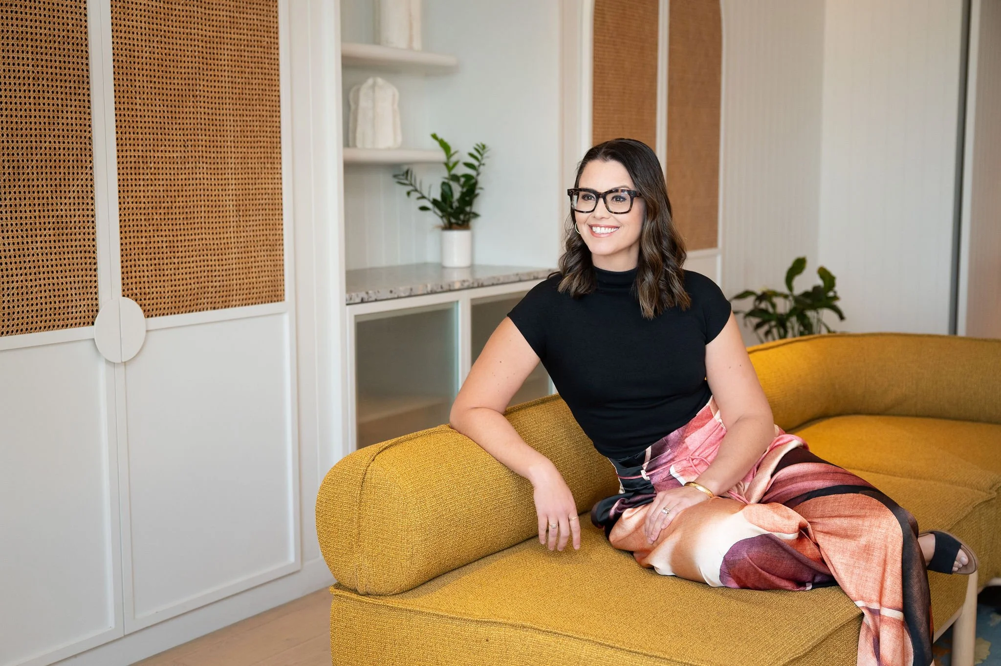 A woman with dark hair, glasses, and a black top sitting on a yellow sofa, smiling in a modern room with white and wood-accented walls and houseplants.