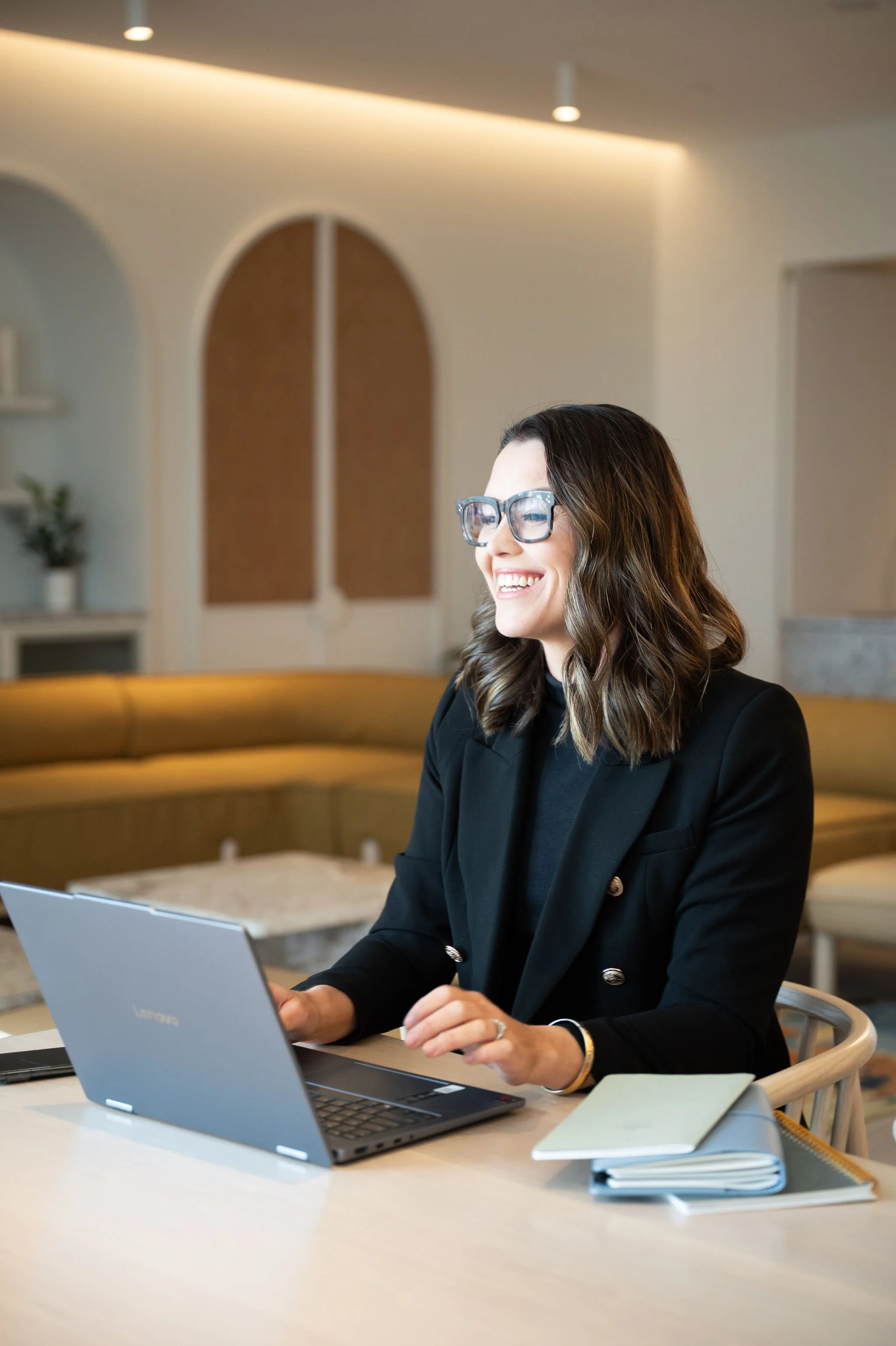 A woman with glasses and wavy brown hair working on a laptop at a desk, smiling.
