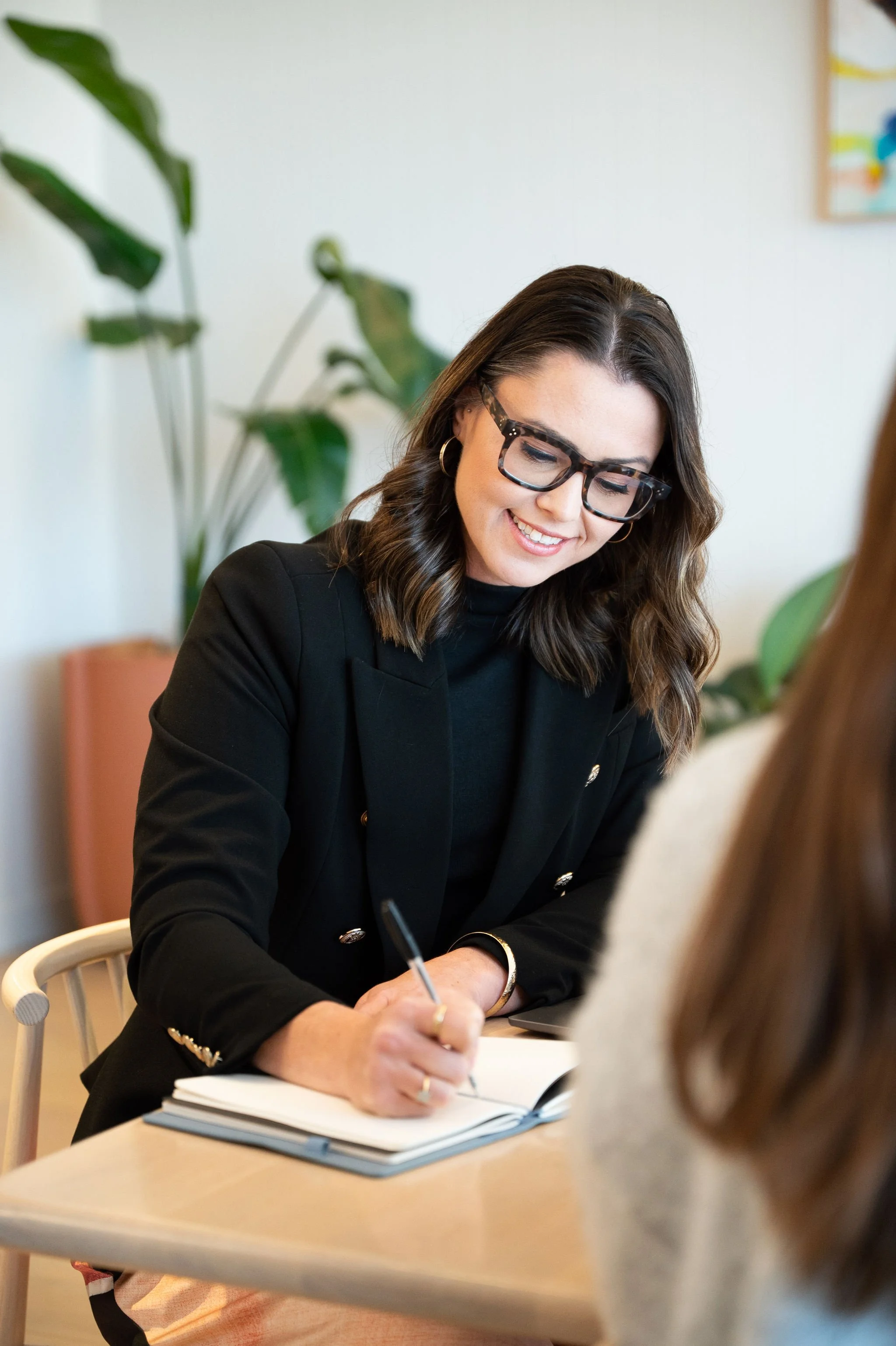 A woman with long dark hair, wearing glasses and a black blazer, sitting at a table and writing in a notebook during a meeting or discussion.