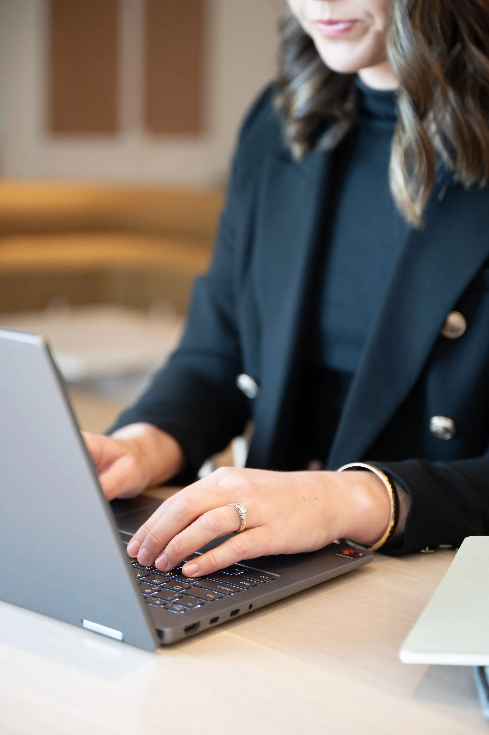 A woman wearing a black blazer and jewelry is working on a laptop at a desk.