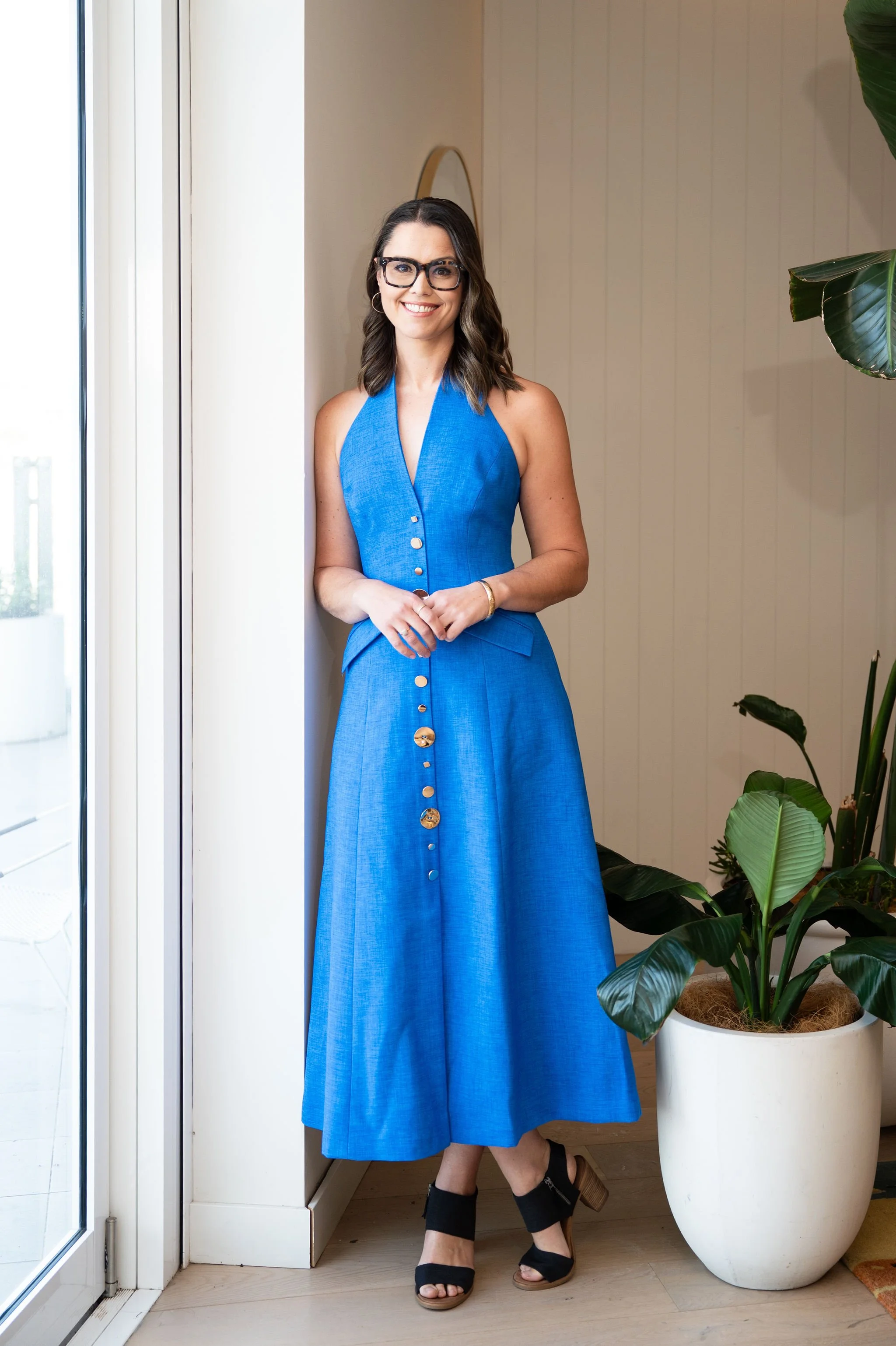 A woman wearing glasses and a blue dress stands indoors next to a large potted green plant near glass doors, smiling at the camera.