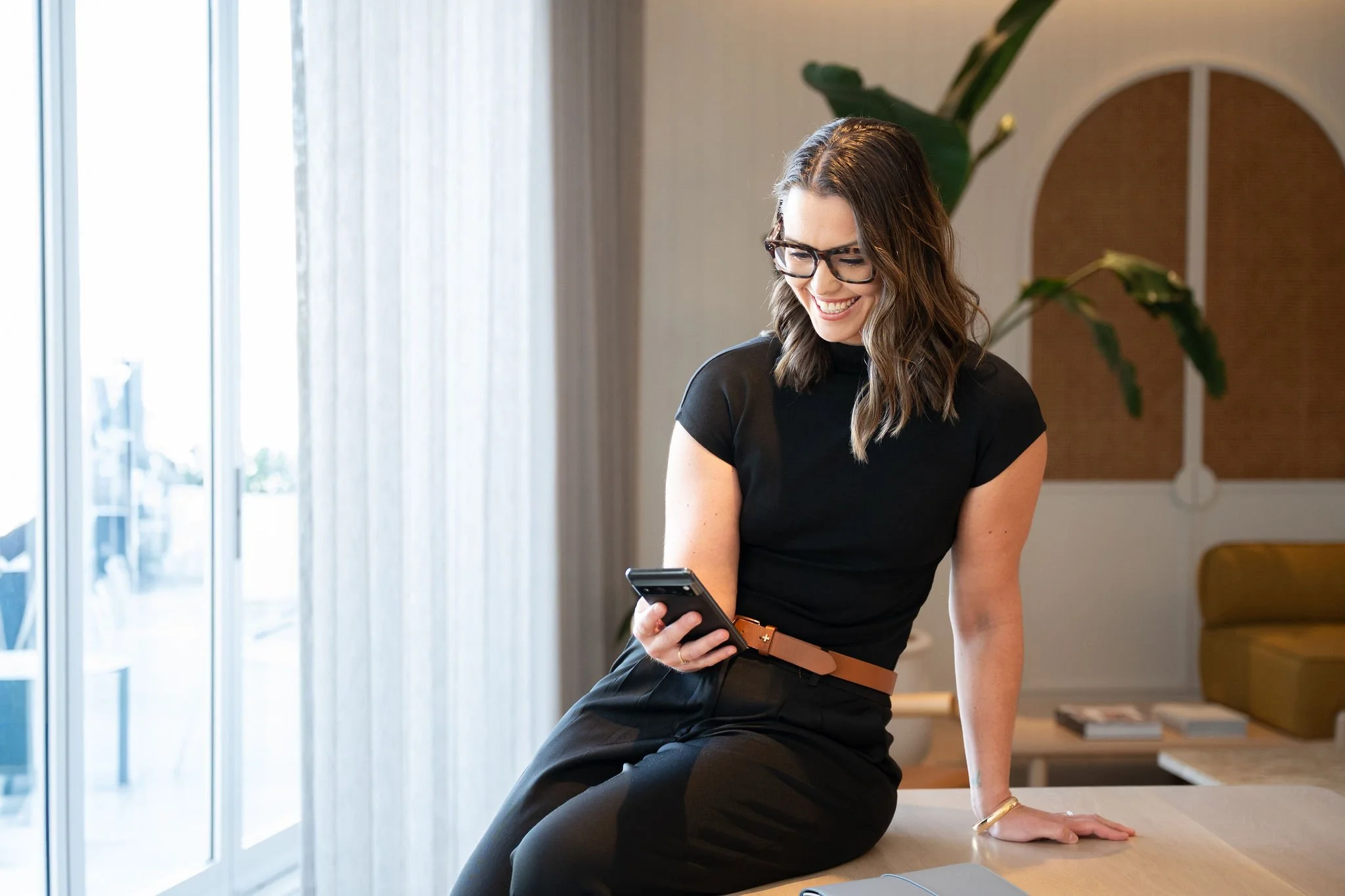A woman with wavy brown hair and glasses, wearing a black top and black pants, sitting on a kitchen counter, smiling while looking at her phone. The background includes a large window with sheer curtains, a potted plant, and modern kitchen decor.