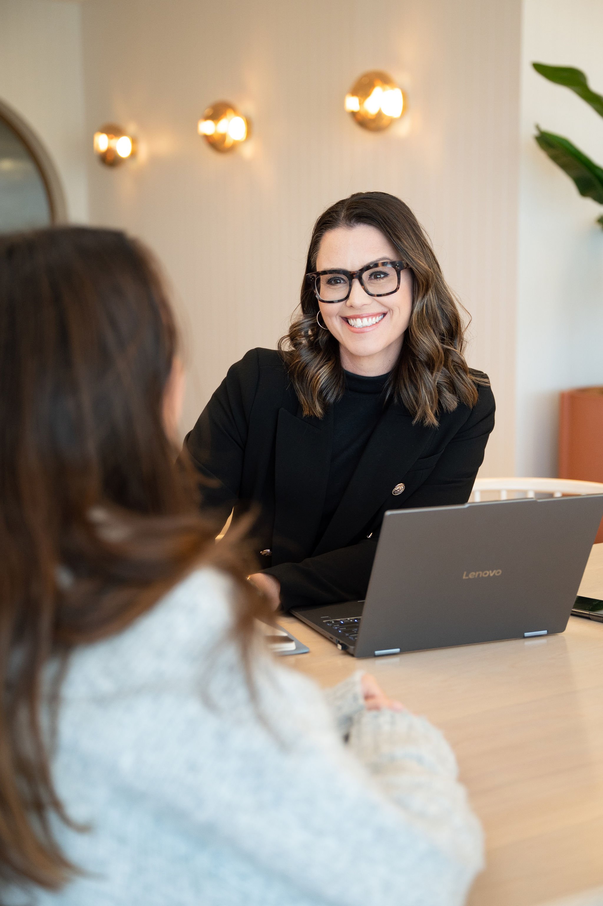A woman with glasses and wavy brown hair smiling during a conversation at a meeting, with a laptop open in front of her, in a modern office setting with warm lighting and a plant in the background.