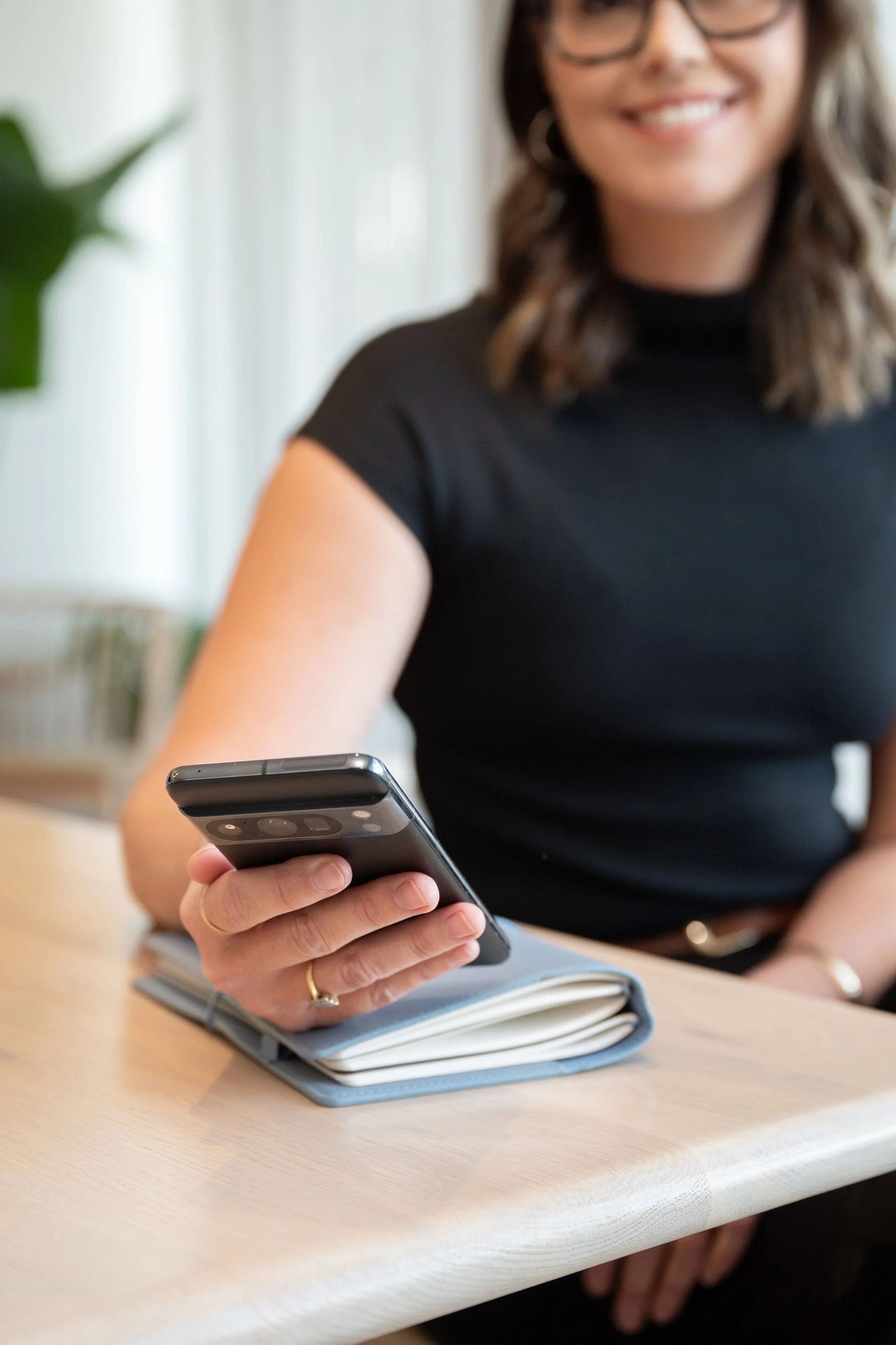 A woman sitting at a table holding a smartphone in her right hand. She is wearing a black shirt, gold rings, and glasses. There is a blue notebook with a pen on the table in front of her.
