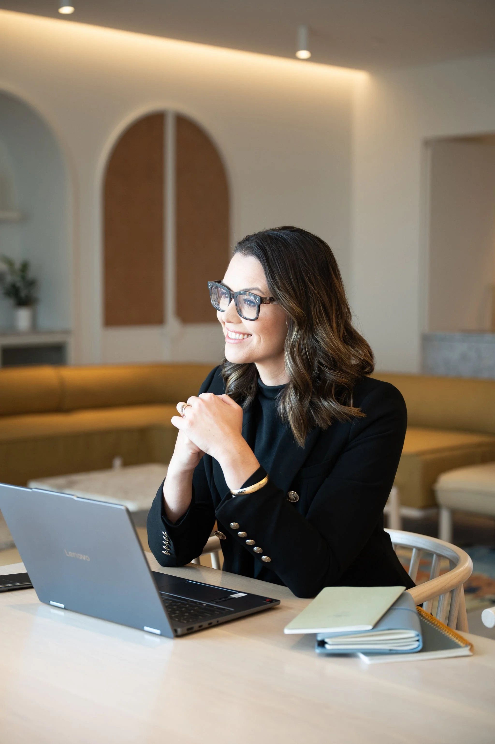 A woman with glasses and wavy brown hair sitting at a table with a laptop, smiling, in a modern, well-lit room.