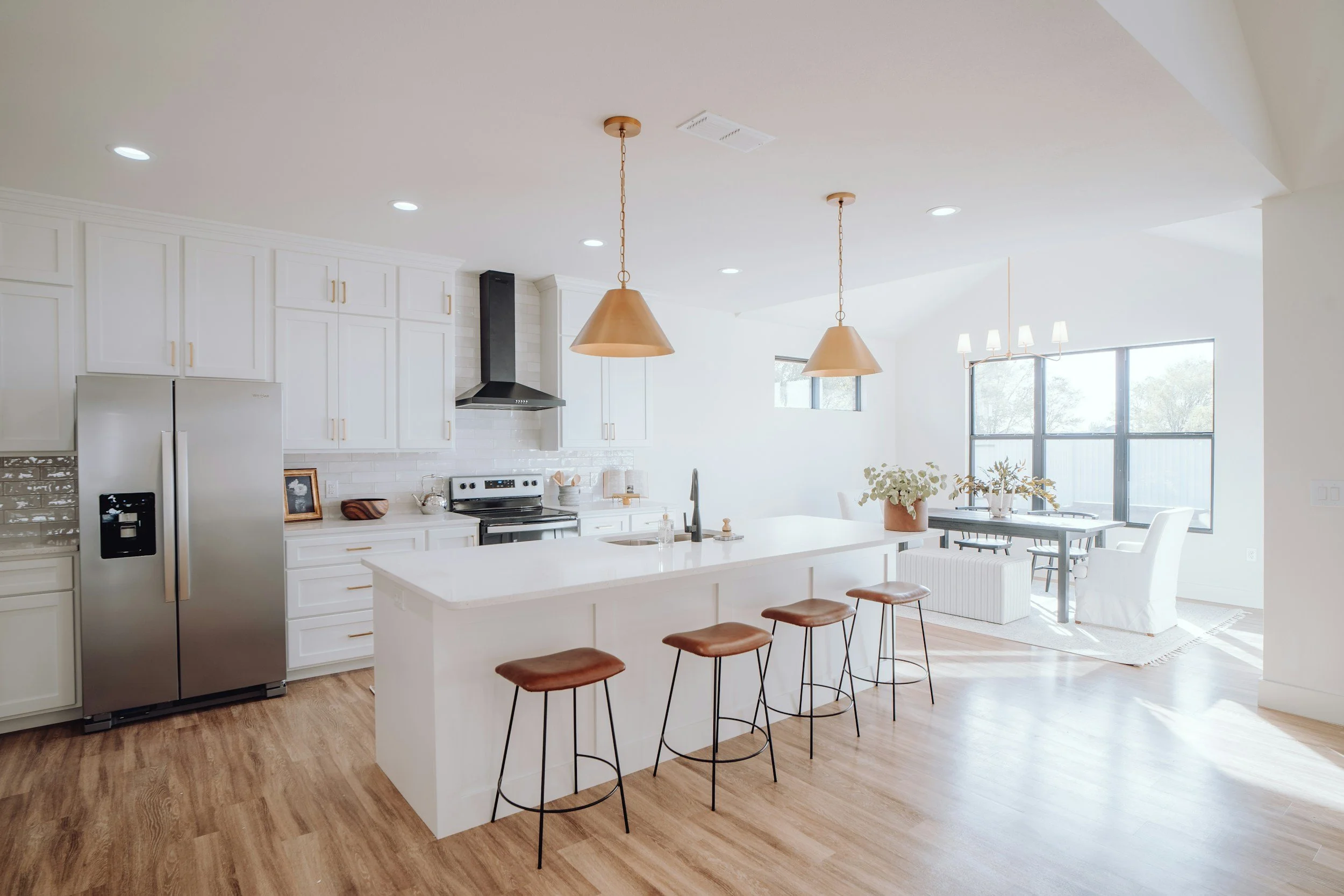 Bright, modern kitchen with white cabinetry, stainless steel refrigerator, black oven and range, light wood flooring, central island with white countertop and four brown stools. Adjacent dining area with large windows, hanging lights, white chairs, and plants.