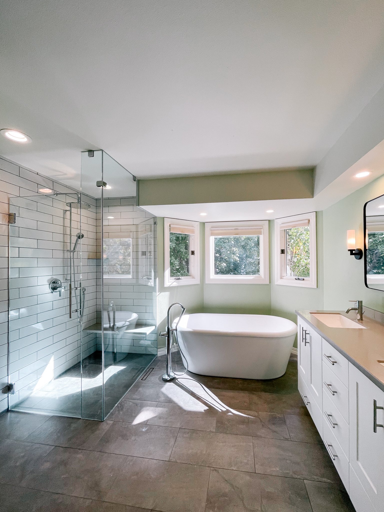 Modern bathroom with glass-enclosed shower, freestanding bathtub, and white vanity with double sinks, illuminated by natural light from three windows.