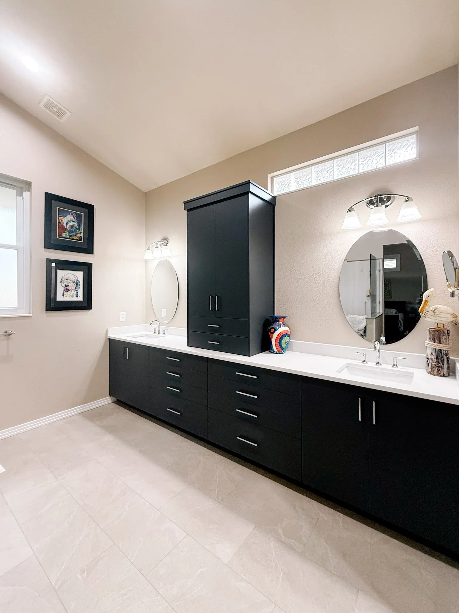 A modern bathroom with a black cabinetry vanity, round mirrors, tan tiled floor, and decorative objects including a colorful vase and a sculpture of a bird.