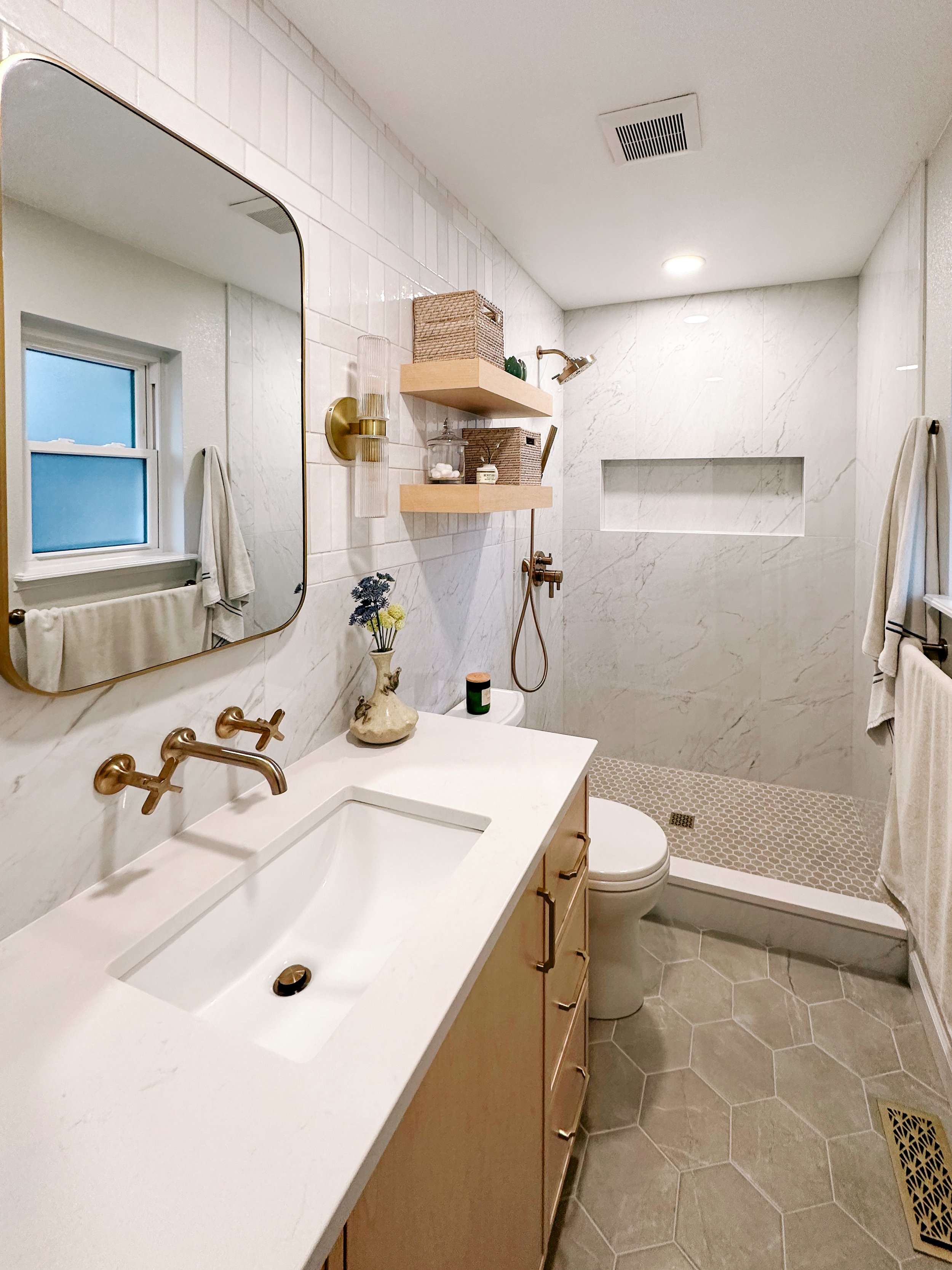 Modern bathroom with white marble walls, vanity with beige cabinet and white countertop, large mirror, brass fixtures, small decorative vase with flowers, shelf with baskets and toiletries, walk-in shower with niche, hexagonal tile flooring, and a wi