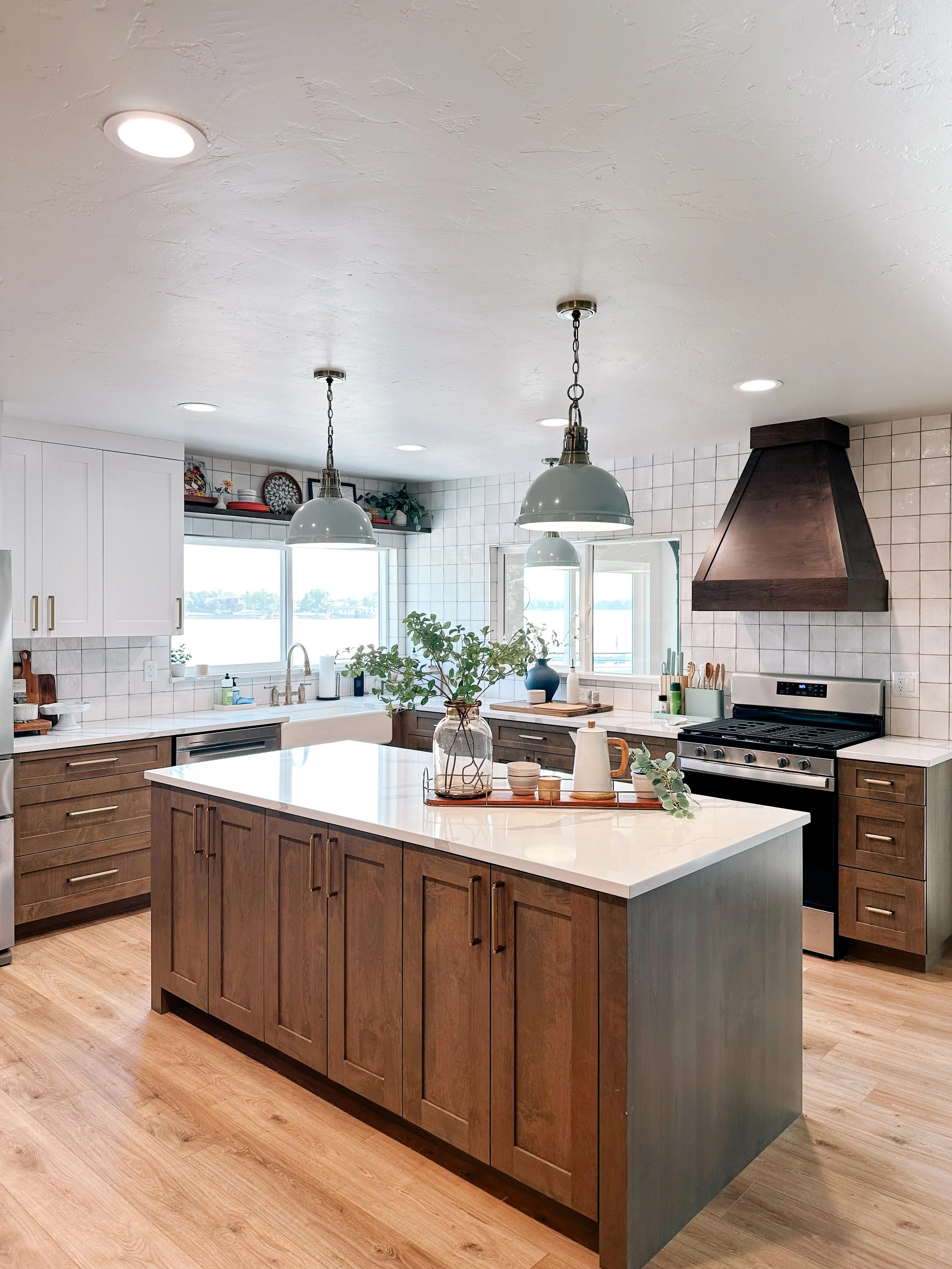 Modern kitchen with white upper cabinets, dark lower cabinets, a large island with a white countertop, stainless steel appliances, a window with outside view, and decorative items like plants and dishes.
