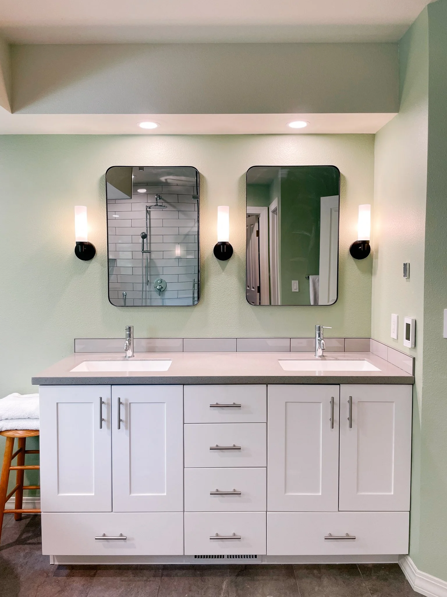 Bathroom vanity with two sinks, white cabinets, two mirrors, and wall-mounted lights, with a shower visible in the mirror reflection.