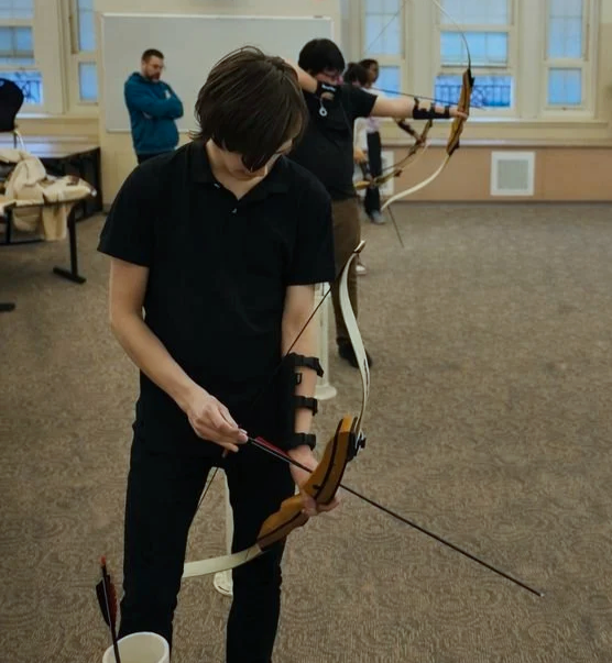 People practicing archery indoors, with one person in the foreground preparing a bow and arrow, and others in the background aiming their bows.