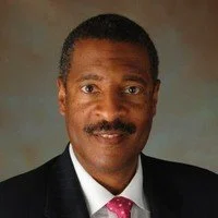 A professional portrait of a middle-aged African American man with short black hair and mustache, wearing a dark suit, white shirt, and pink tie, posing against a neutral background.