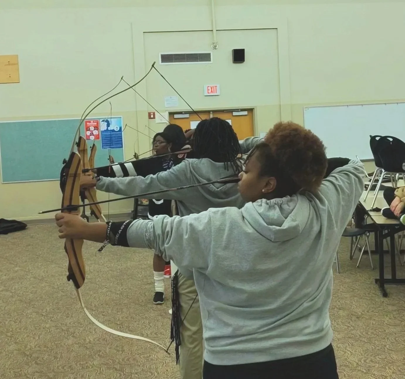 Group of people practicing archery indoors, aiming bows at targets.