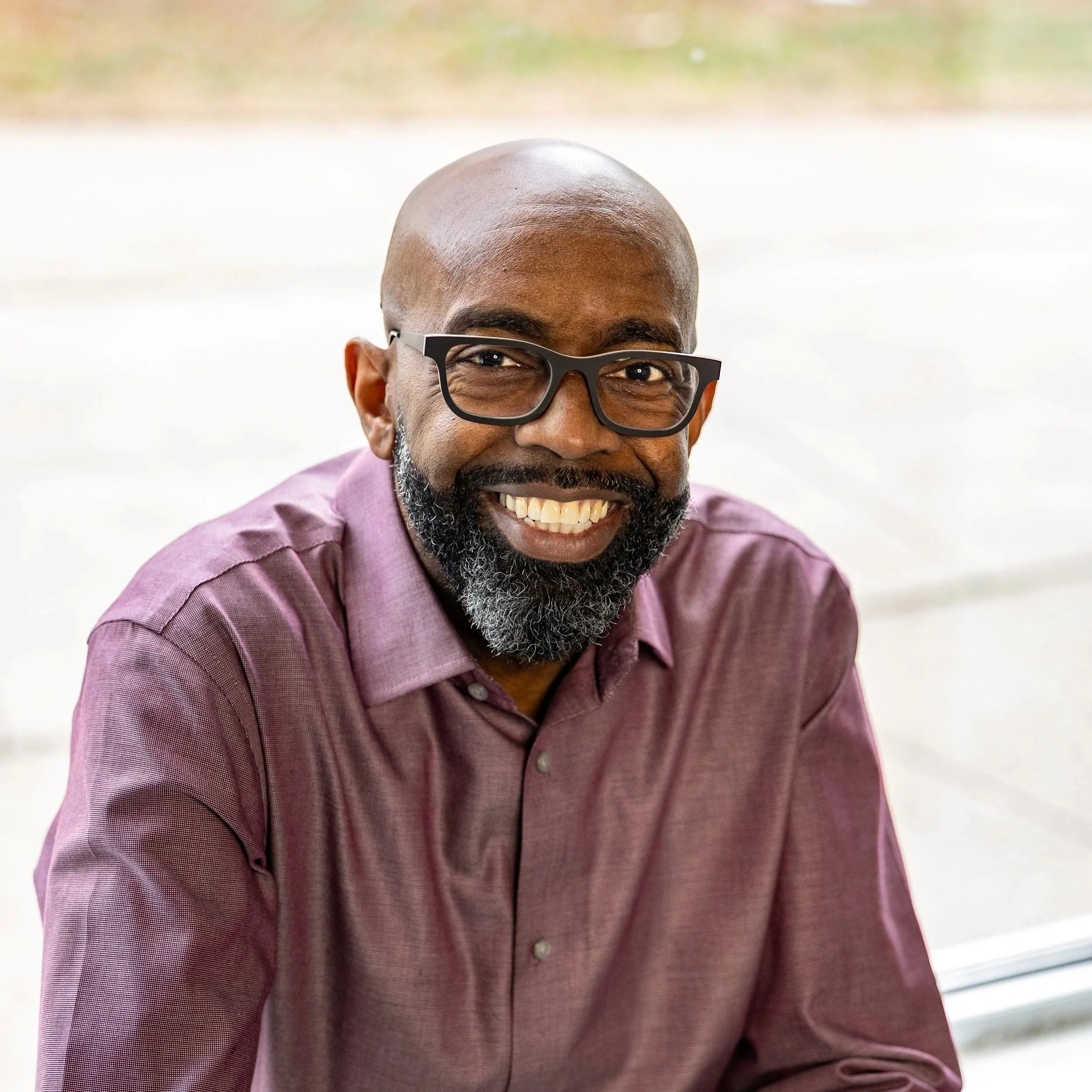 Close-up photo of a smiling Black man with a beard and glasses, wearing a purple shirt, sitting near a large window.
