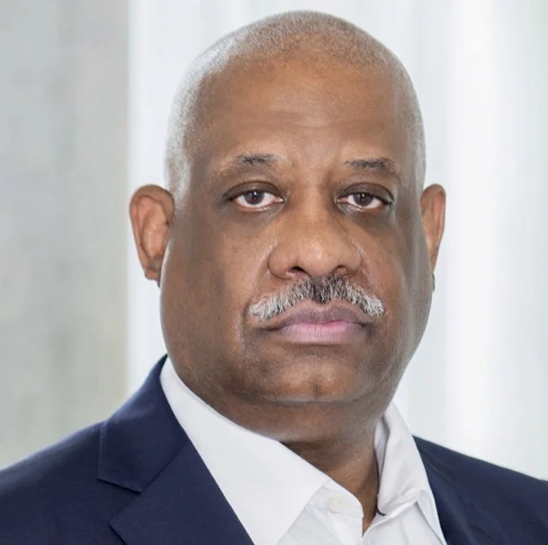 A close-up portrait of a middle-aged man with a serious expression, wearing a navy suit and white shirt, with a light background.