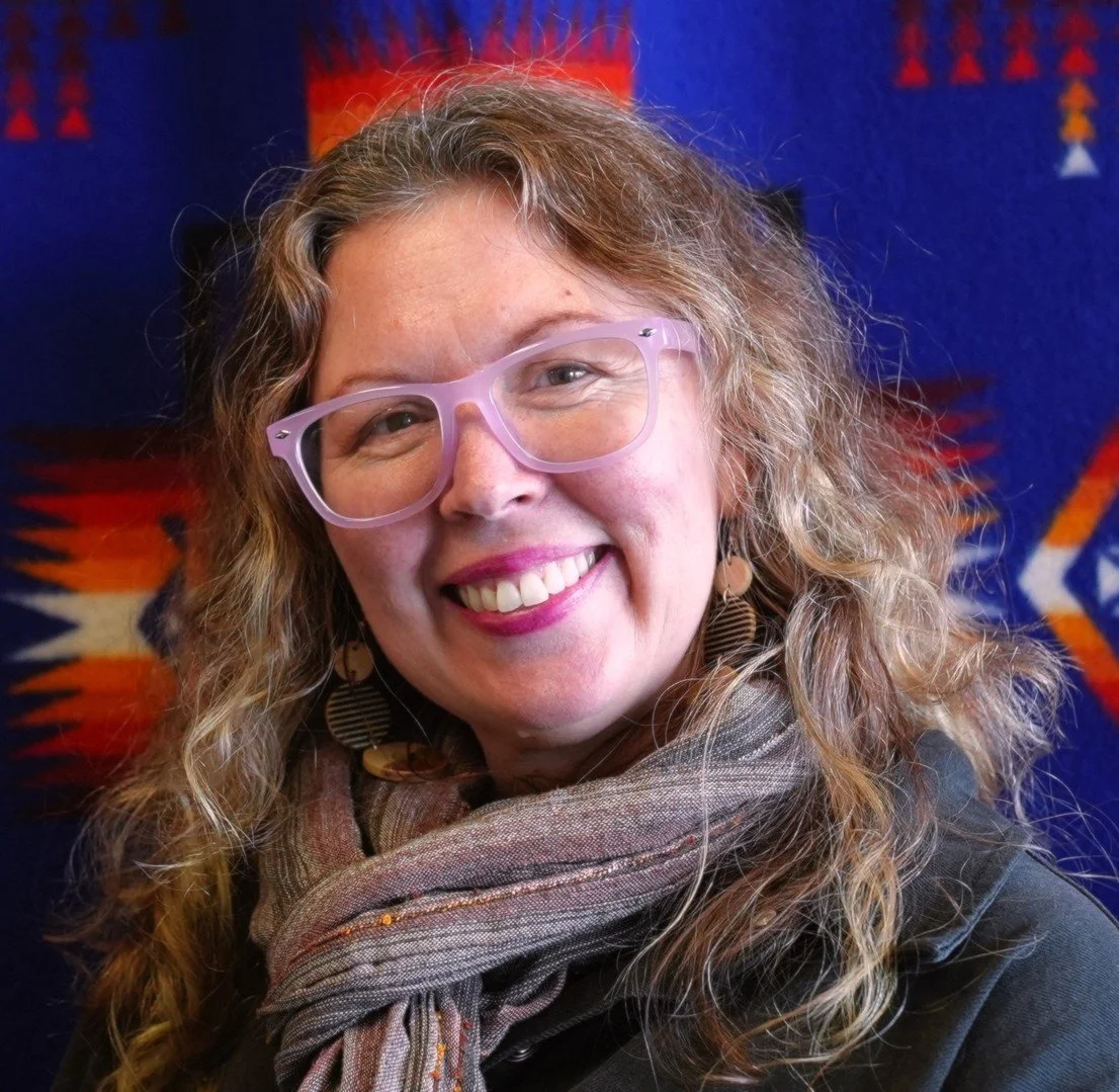 A woman with curly blonde hair and pink glasses smiles in front of a colorful textile background, wearing earrings and a scarf.
