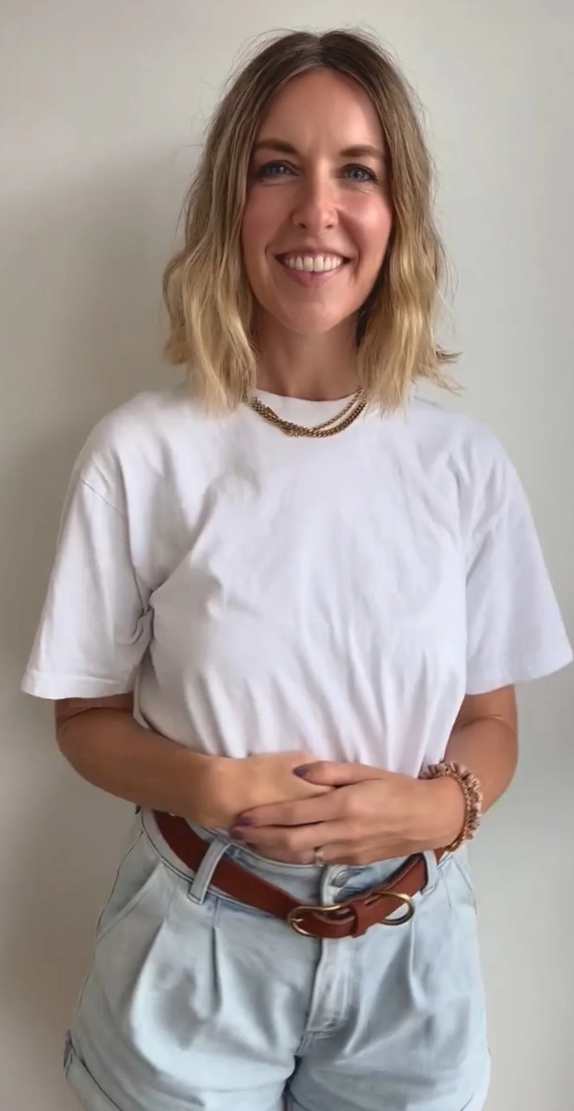 A woman with shoulder-length wavy blonde hair is smiling and standing against a plain light-colored wall. She is wearing a white t-shirt, a layered gold chain necklace, a brown belt, and light-colored pants.