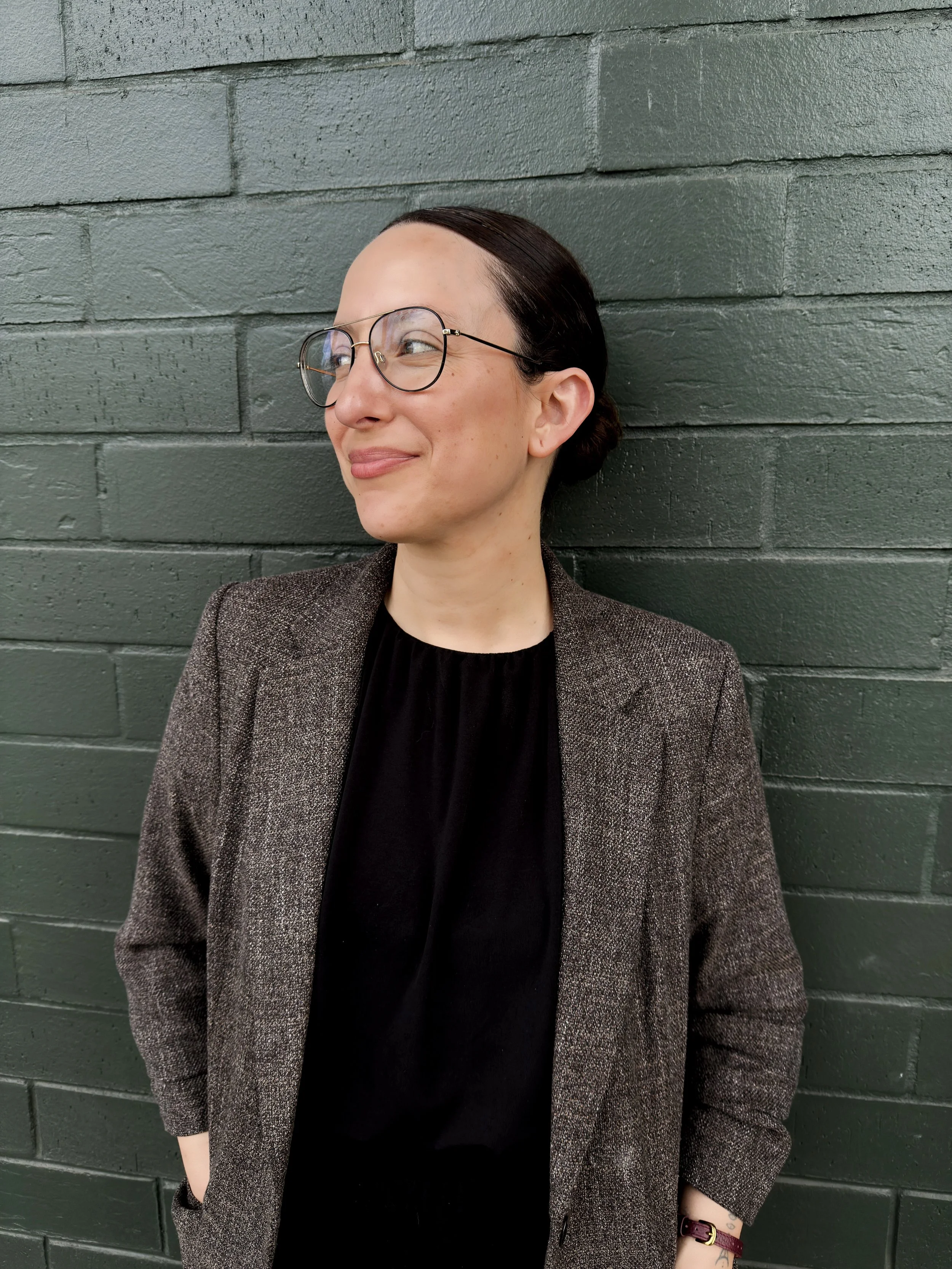 A woman with dark hair pulled back, wearing glasses, a black top, a gray blazer, and a maroon watch, smiling while leaning against a dark gray brick wall.