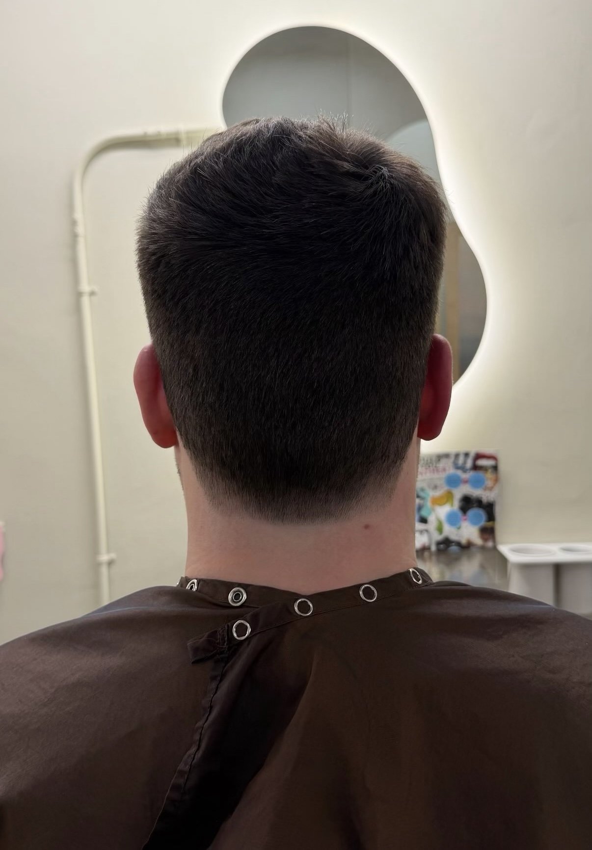 Back of a person's head with short, dark hair, sitting in a salon chair with a brown cape around their shoulders, looking into a mirror.