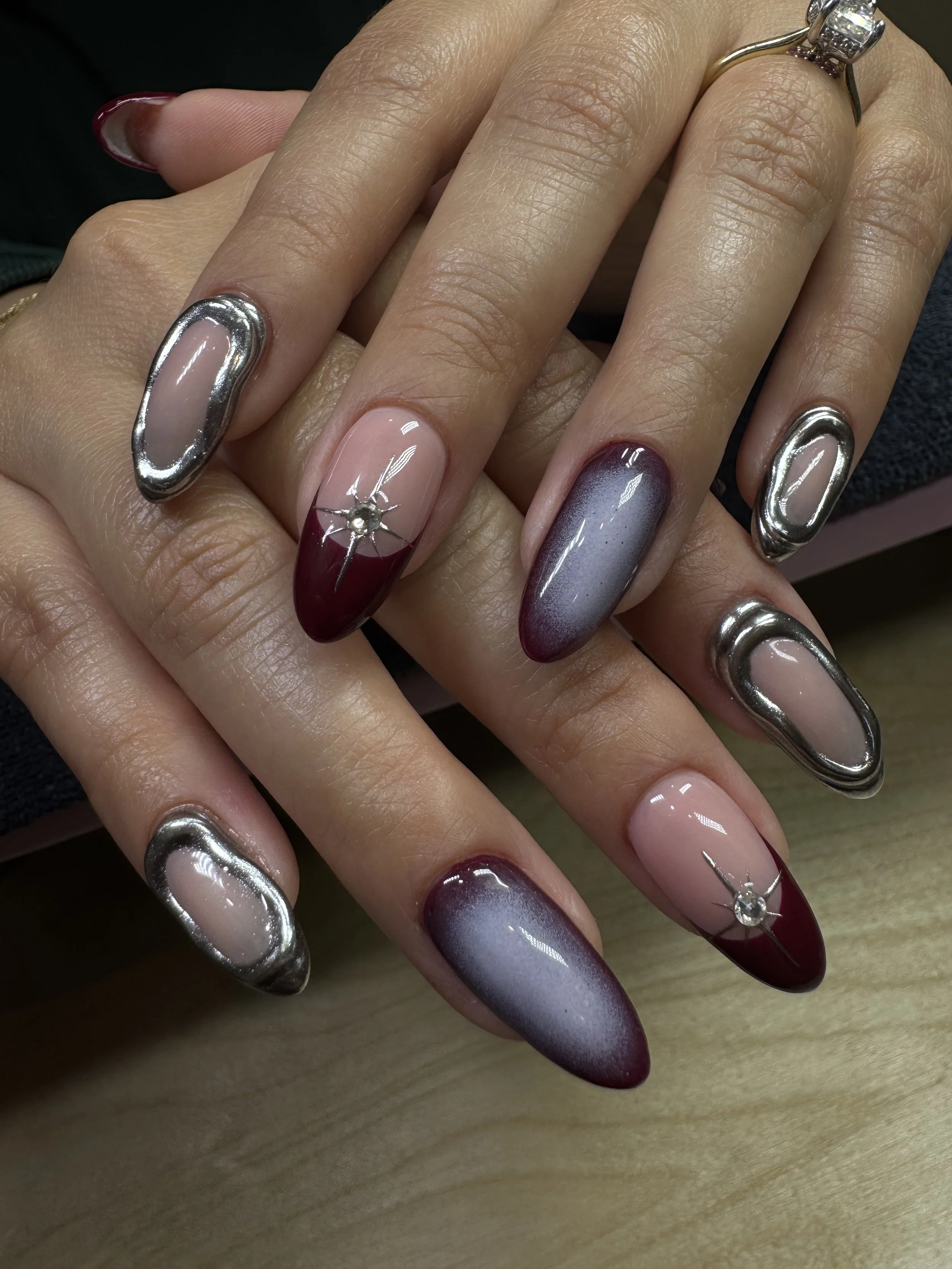 Close-up of a woman's hands with manicured nails in an array of colors and designs, including metallic silver, deep red with rhinestone embellishments, and gradient shades of purple and gray.