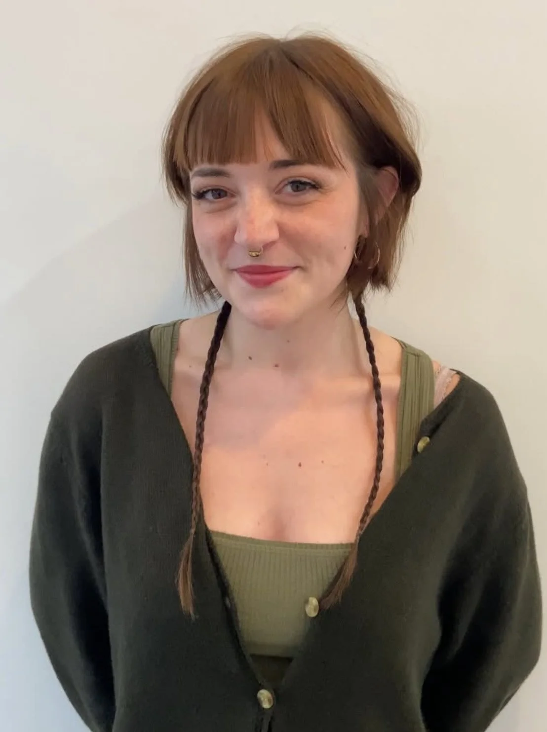 A woman with short, reddish-brown hair, wearing a septum ring, black cardigan, and green top, standing against a plain white wall.
