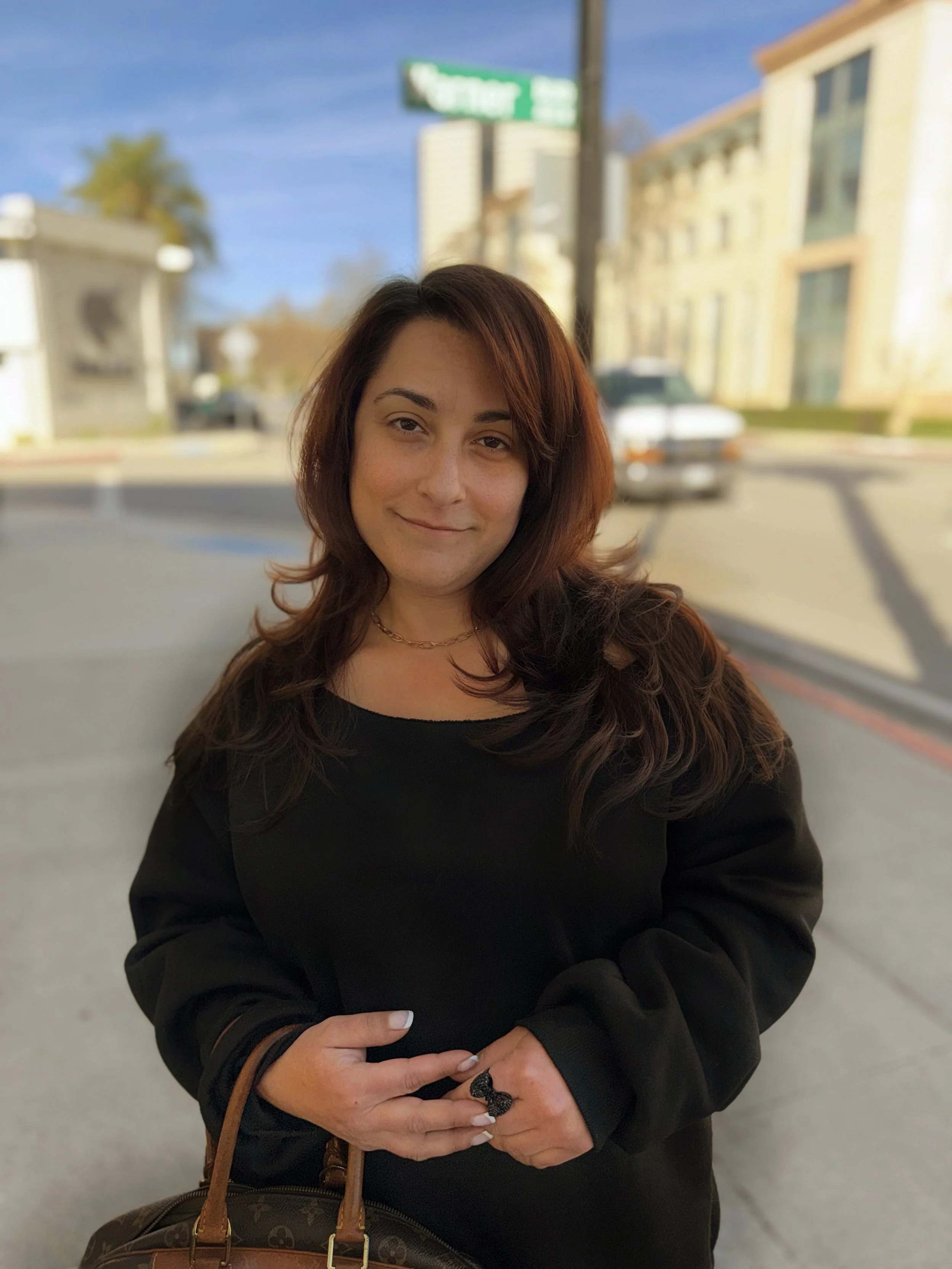 A woman with long brown hair and a black sweater, holding a bag, standing outside on a sunny day with buildings and a street sign in the background.