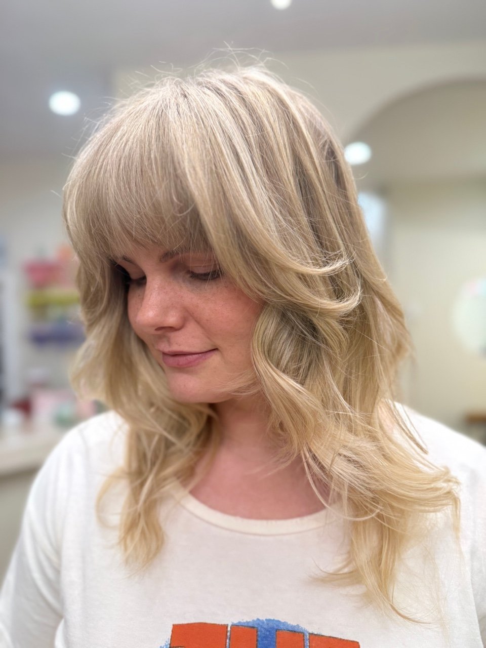 Woman with blonde, wavy hair and bangs, wearing a white T-shirt with colorful graphics, in a salon or similar setting.