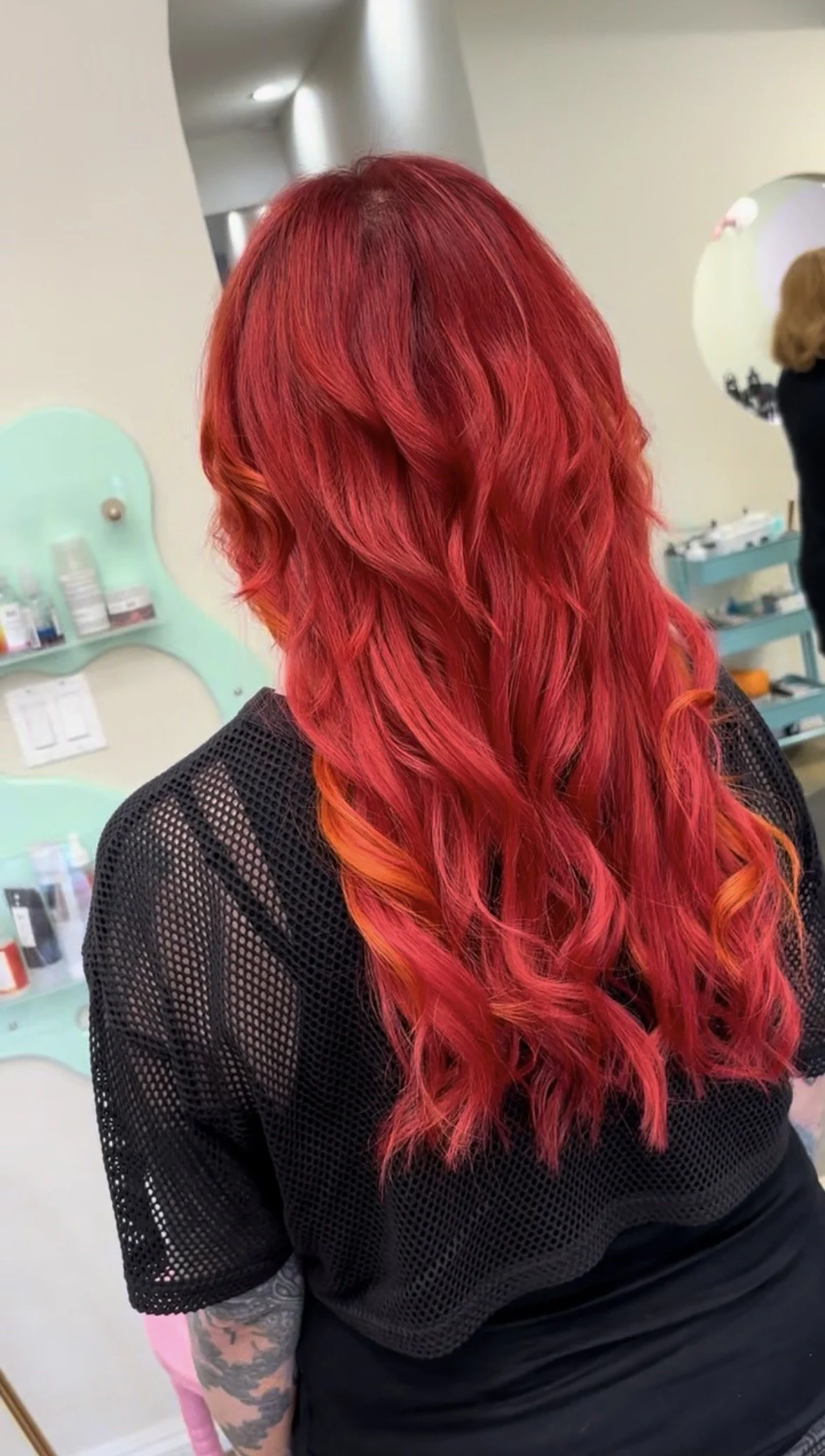 A person with long, vibrant red hair styled in loose waves, wearing a black mesh shirt, standing in a salon or studio with shelves of hair products in the background.