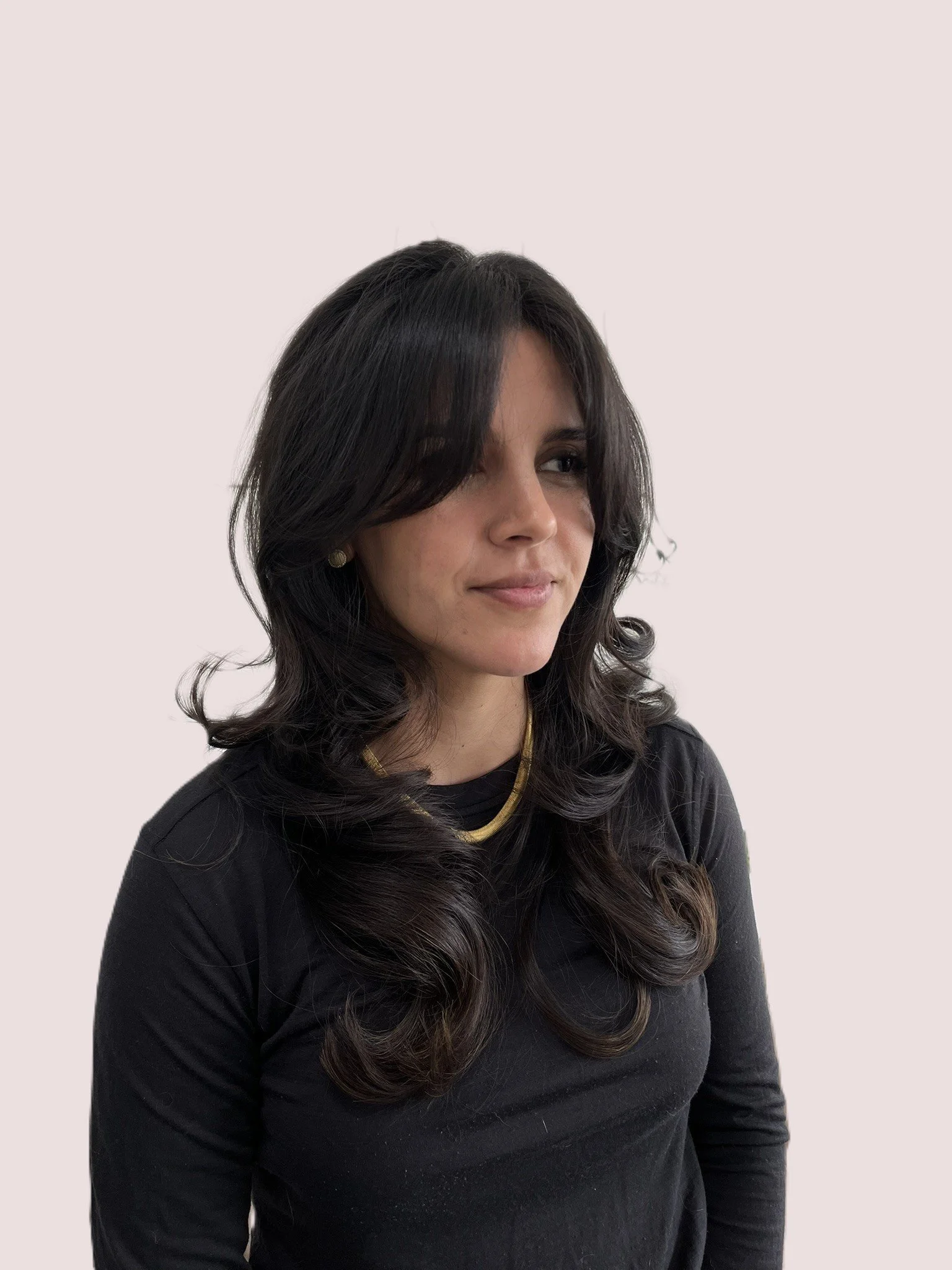 A woman with long, dark, wavy hair wearing a black top, gold necklace, and small earrings, looking slightly to her left, against a plain light-colored background.