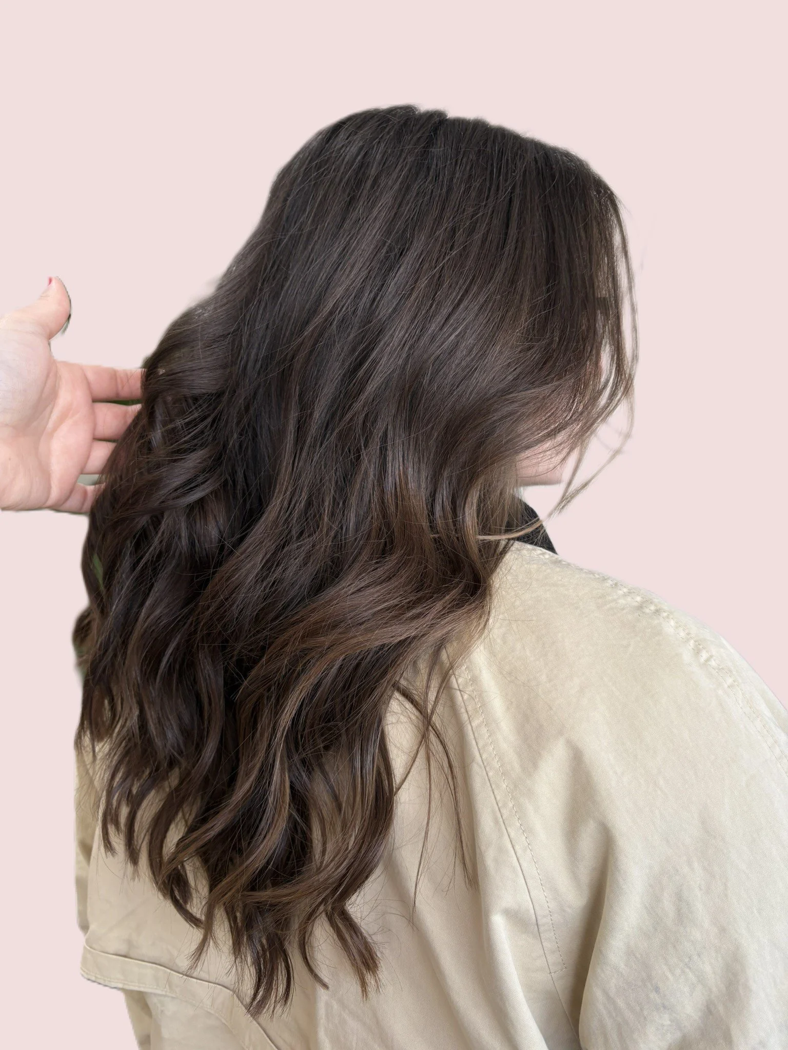 A woman with long, wavy, dark brown hair wearing a beige shirt, looking down with her back to the camera, against a light pink background.