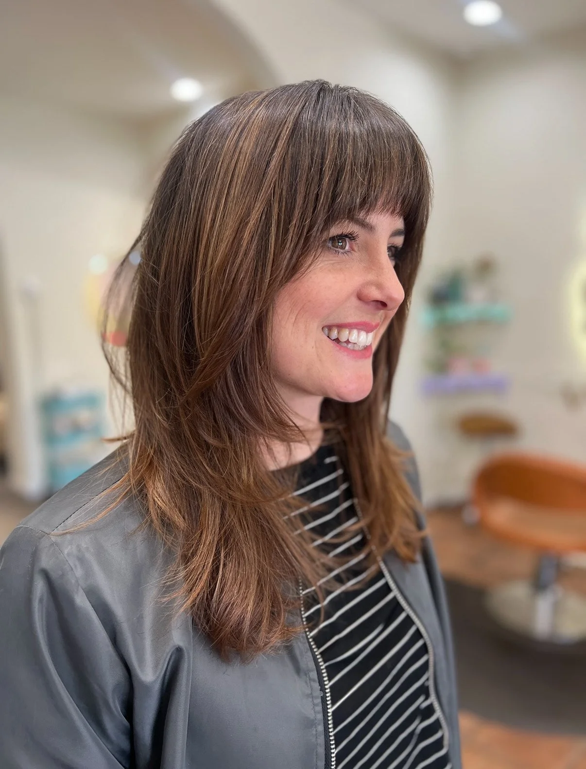 A woman with brown, layered hair and bangs, smiling and looking to the right. She is wearing a black and white striped shirt and a gray jacket. The background is blurred, showing a room with light-colored walls and some furniture.