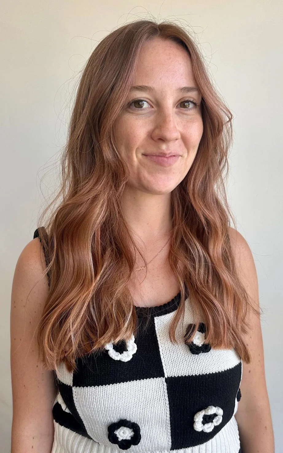 A woman with long wavy auburn hair, fair skin, and light-colored eyes, smiling slightly, wearing a black and white checkered sleeveless top with decorative circular black and white accents, and a delicate necklace.