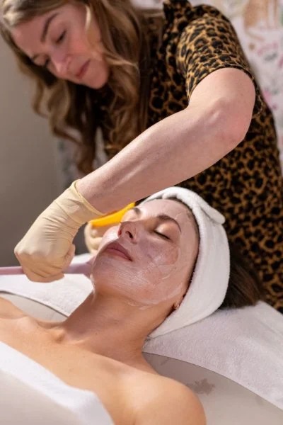 A woman in a leopard-print shirt applies a facial treatment to a woman lying down with a towel wrapped around her head.