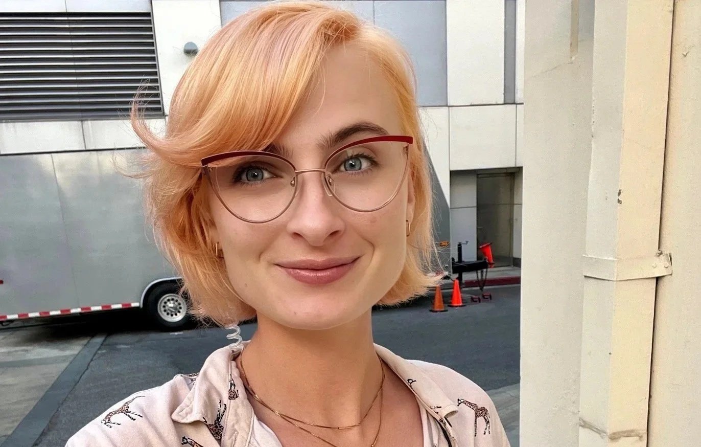 A young woman with short peach-colored hair, wearing glasses and a light-colored jacket decorated with giraffe prints, standing outdoors in front of a building with a parked trailer and traffic cones.
