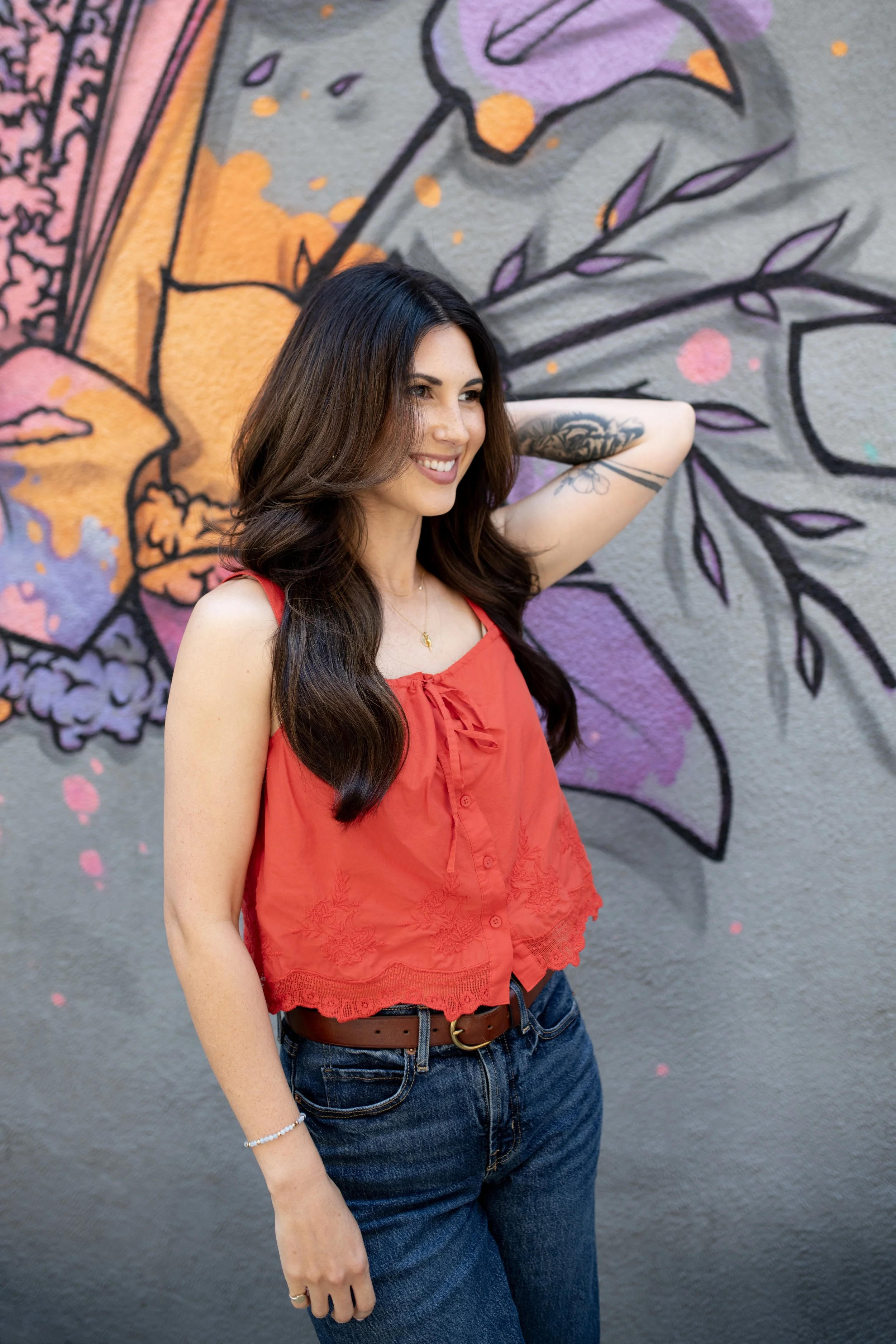 A young woman with long dark hair and tattoos on her arm, smiling and standing with one hand resting on her neck, against a colorful mural of a butterfly on a gray wall. She is wearing a red sleeveless top and blue jeans.