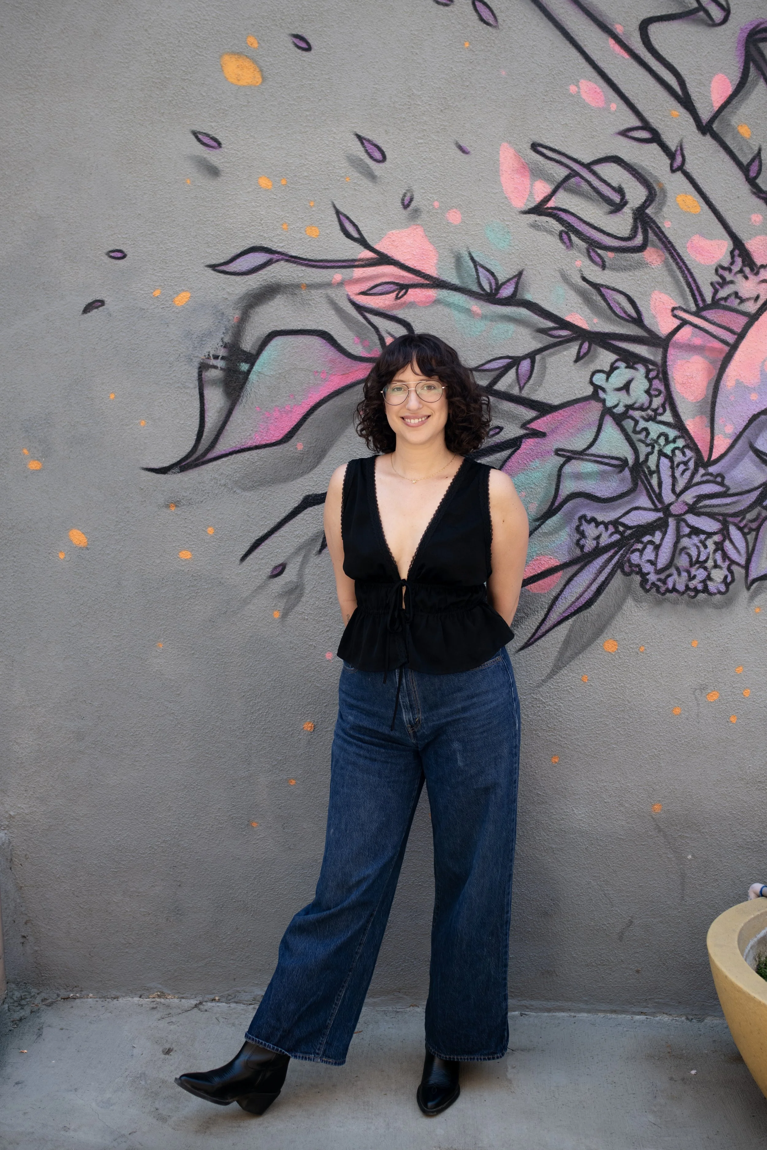 A woman with curly dark hair, glasses, wearing a black sleeveless top, wide-leg blue jeans, and black boots, standing in front of a mural of abstract floral art on a gray wall.