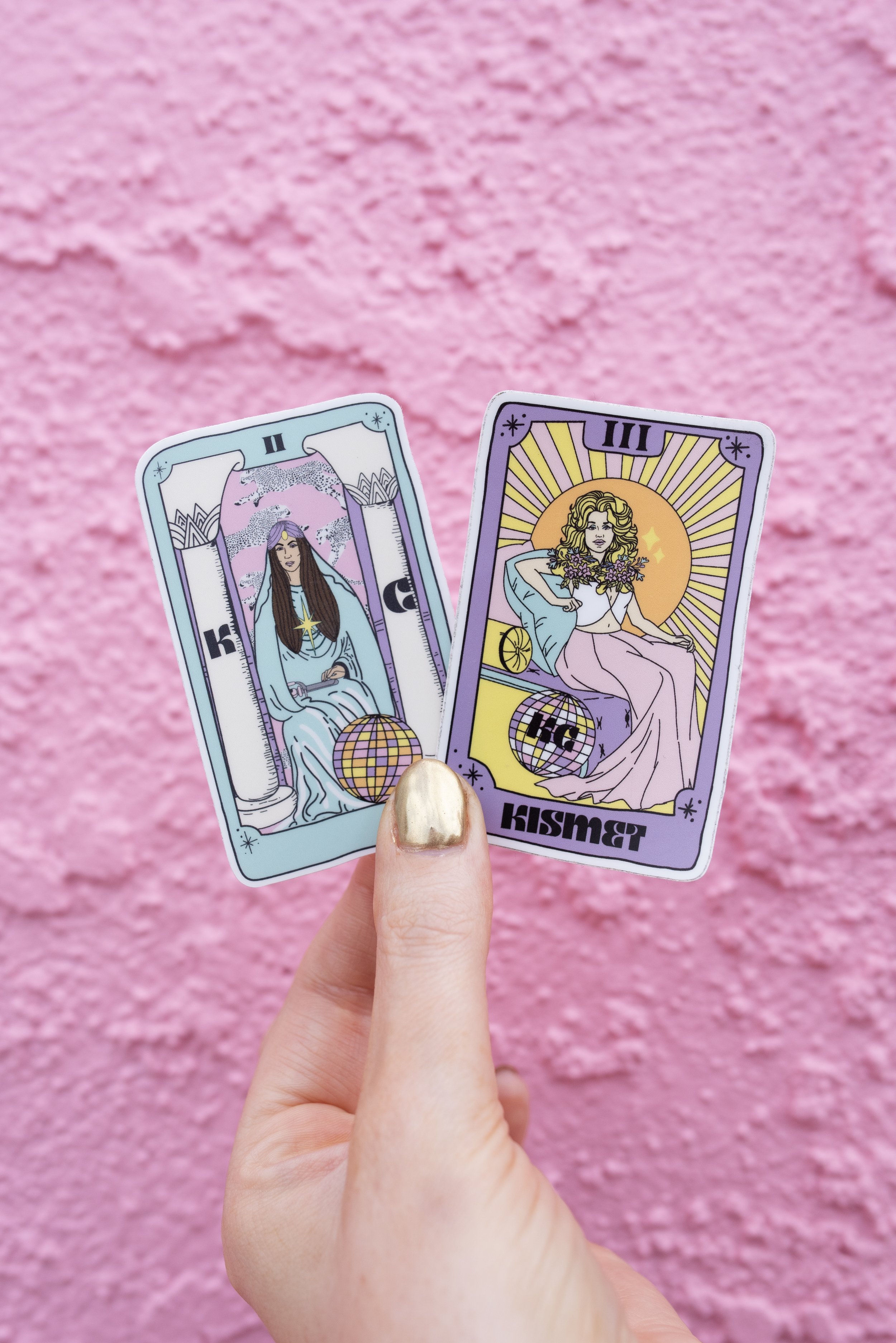 Two tarot cards held by a person's hand in front of a pink textured wall. The left card depicts a woman with long hair, wearing a light blue dress, sitting between two columns with a star above her head, and a globe with a pattern at her feet. The right card features a woman with curly hair, sitting with a bouquet of flowers, with a large sun or moon and rays behind her, and a globe with a pattern at her feet.