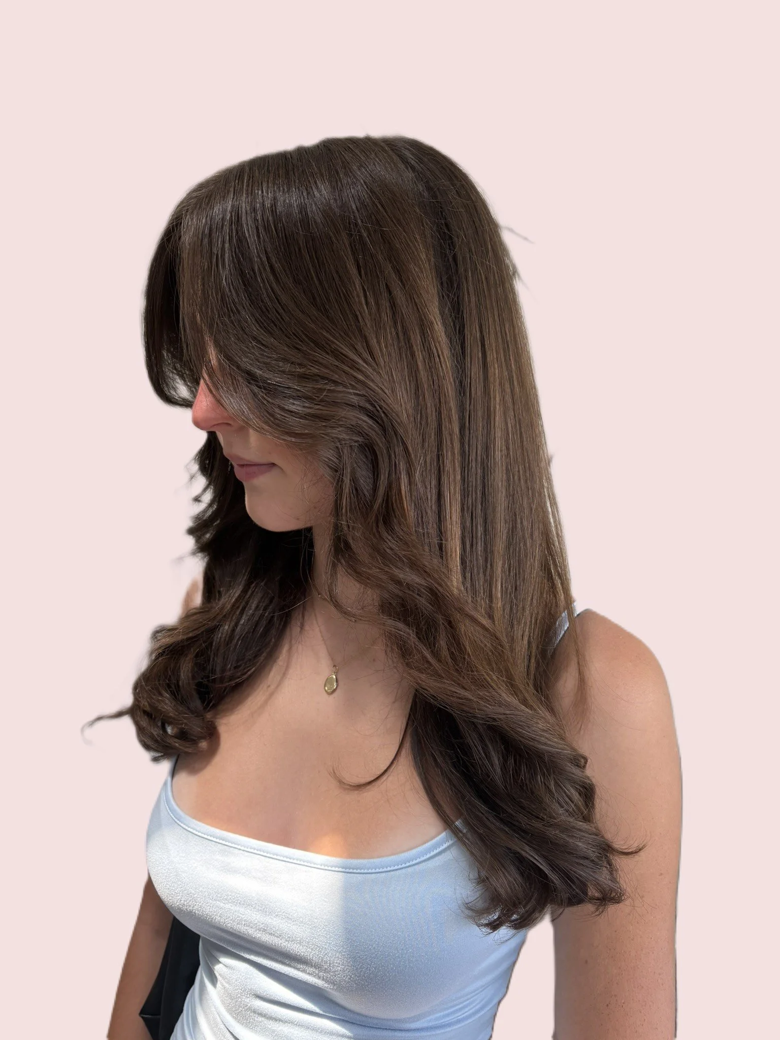 A woman with long, wavy brown hair wearing a white tank top and a gold necklace, with a black bag on her shoulder, against a light pink background.