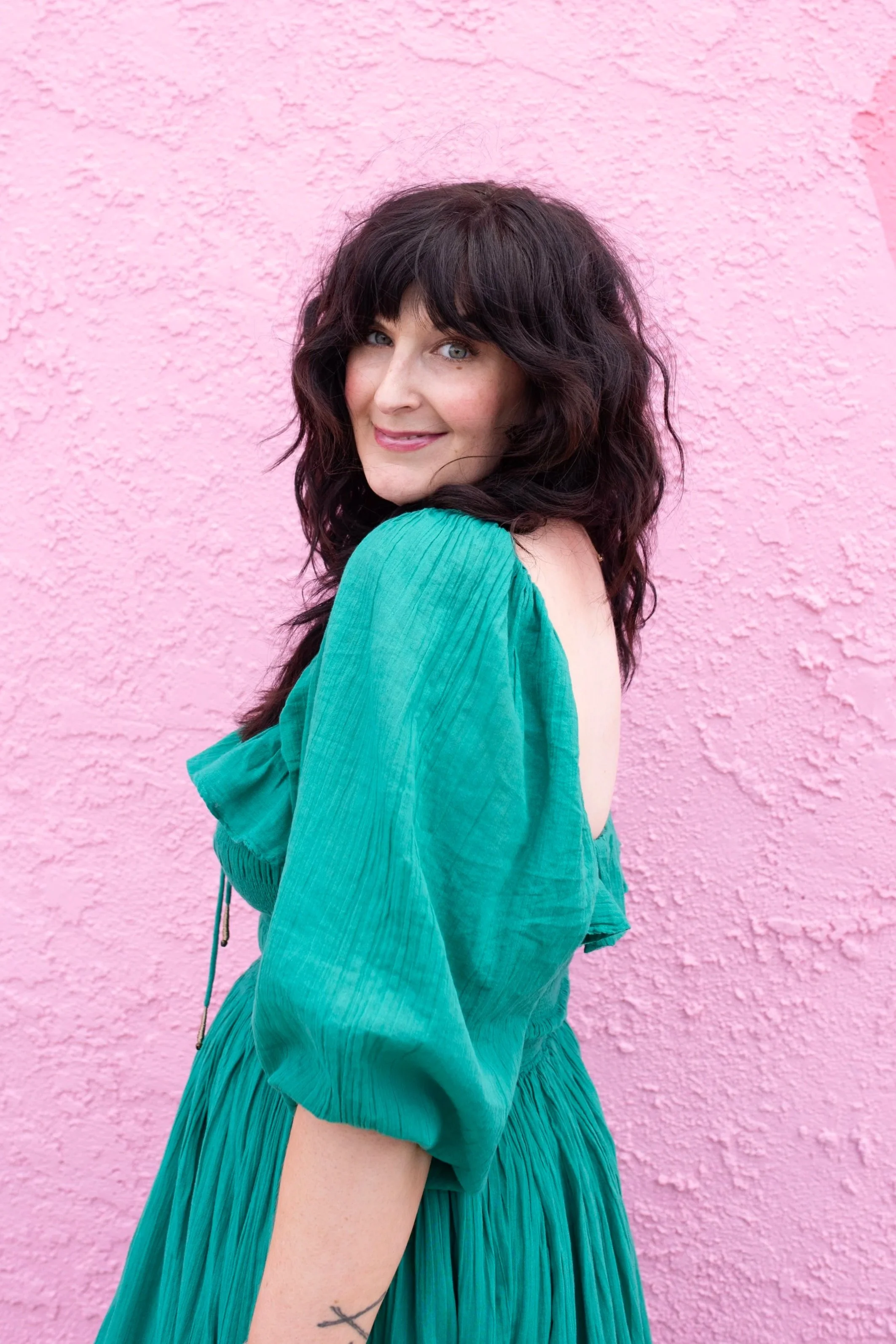 Roxanne Schrieber, with dark wavy hair and blue eyes, smiling, wearing a teal dress with puffy sleeves, standing against a pink textured wall.