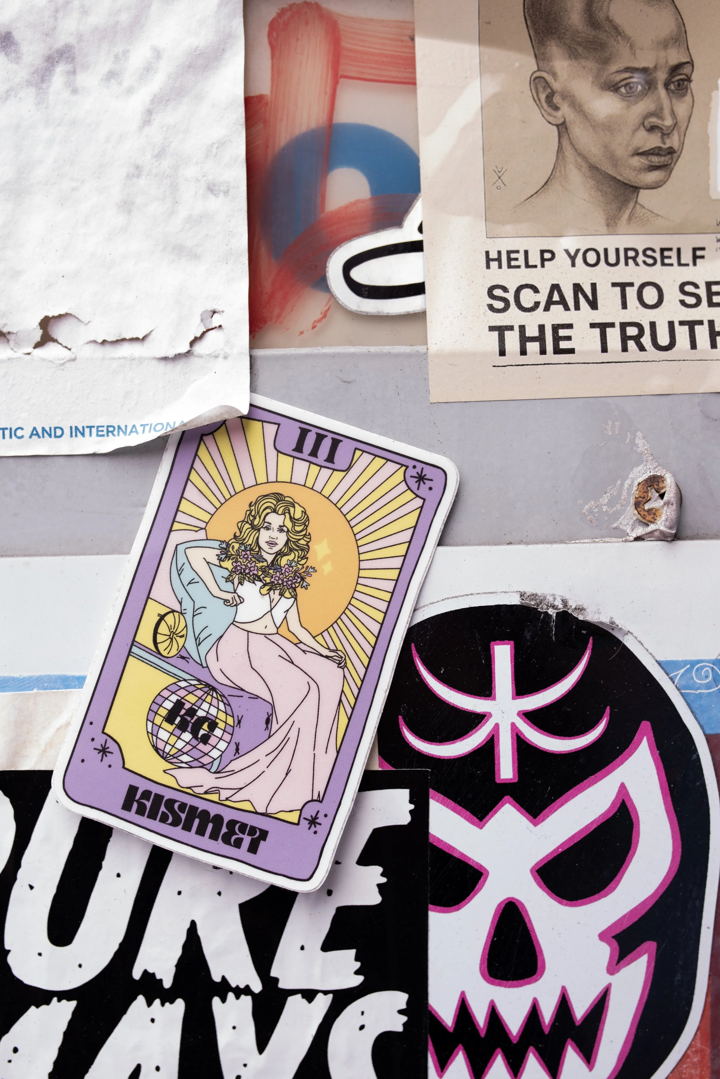 Collection of stickers and posters on a wall, including a tarot card-style illustration of a woman, a skull design with pink outlines, and various torn paper edges.