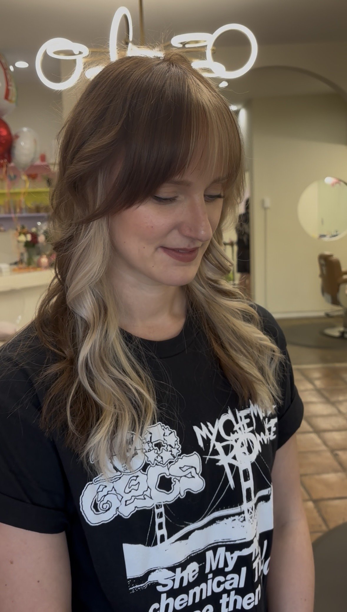 A woman with wavy, shoulder-length hair with blonde highlights, wearing a black T-shirt with white graphic text, smiling slightly while looking down in a salon or hair studio. The background features salon lighting and decorating balloons.