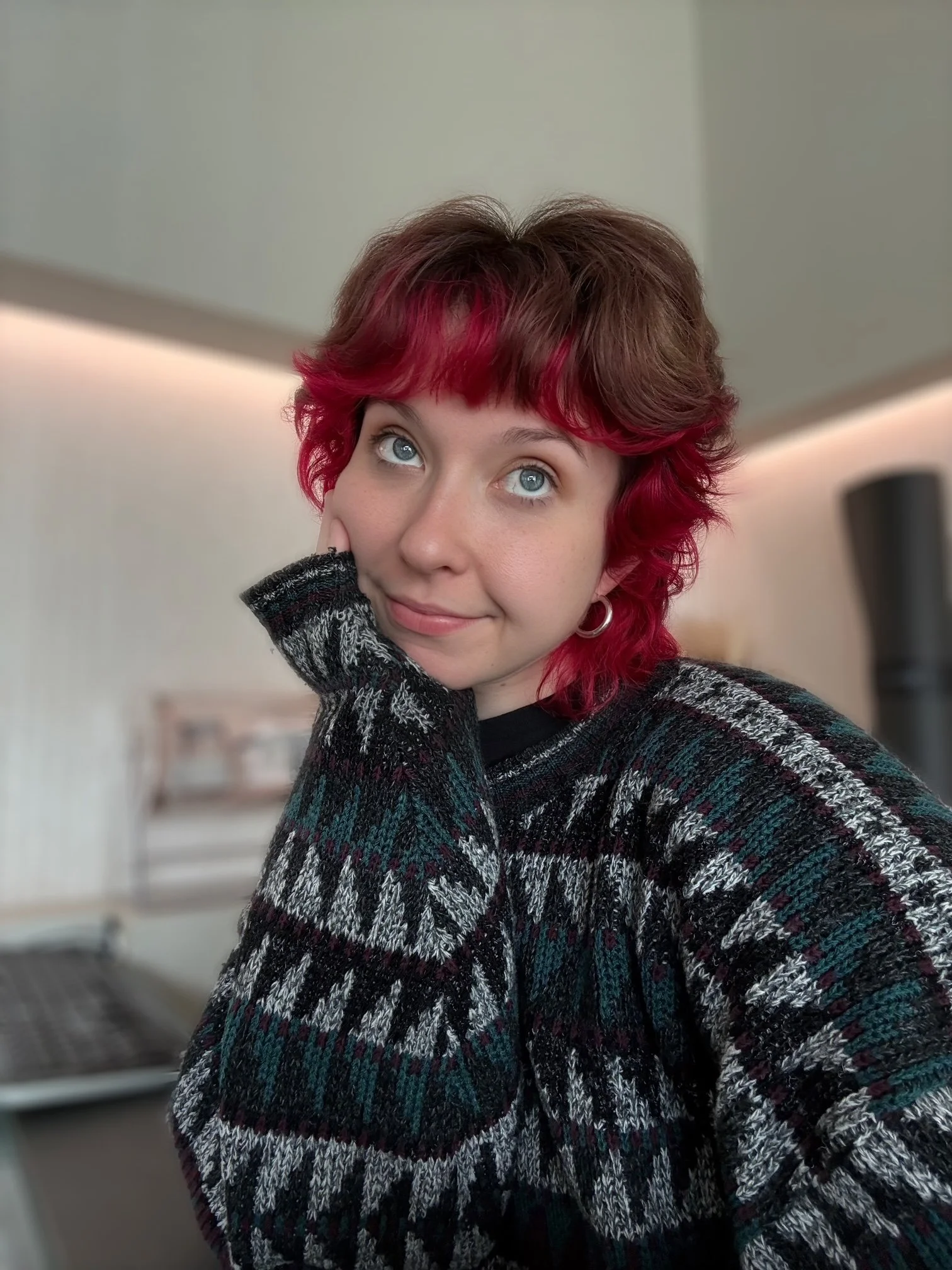 A young woman with short, curly hair with red highlights, wearing a black and gray patterned sweater, looking up with a slight smile, resting her face on her left hand, in an indoor setting with blurred background.