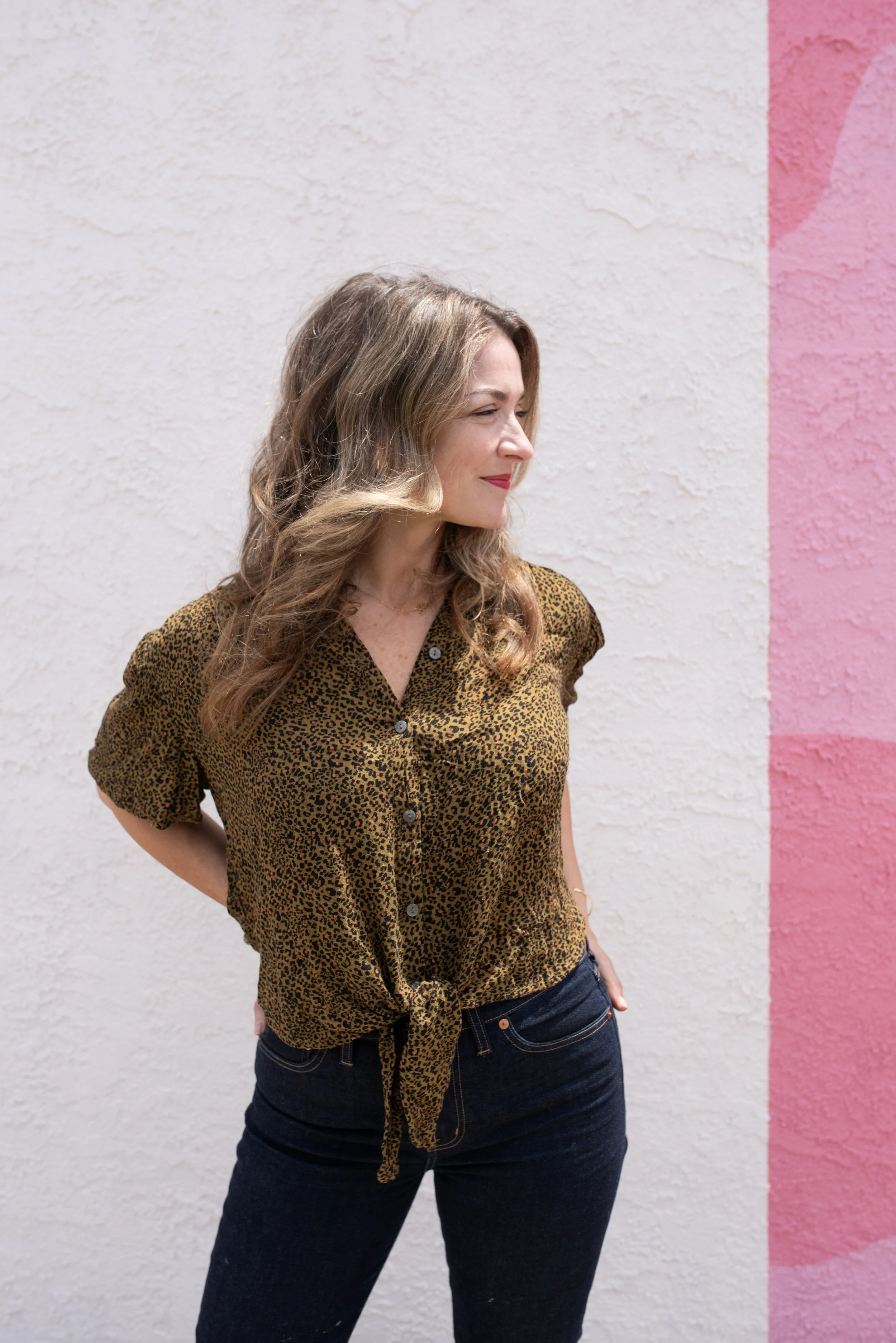 A woman with wavy, shoulder-length hair stands against a wall painted with pink and white sections. She wears a leopard print shirt tied at the waist and dark jeans, looking to her right with a gentle smile.