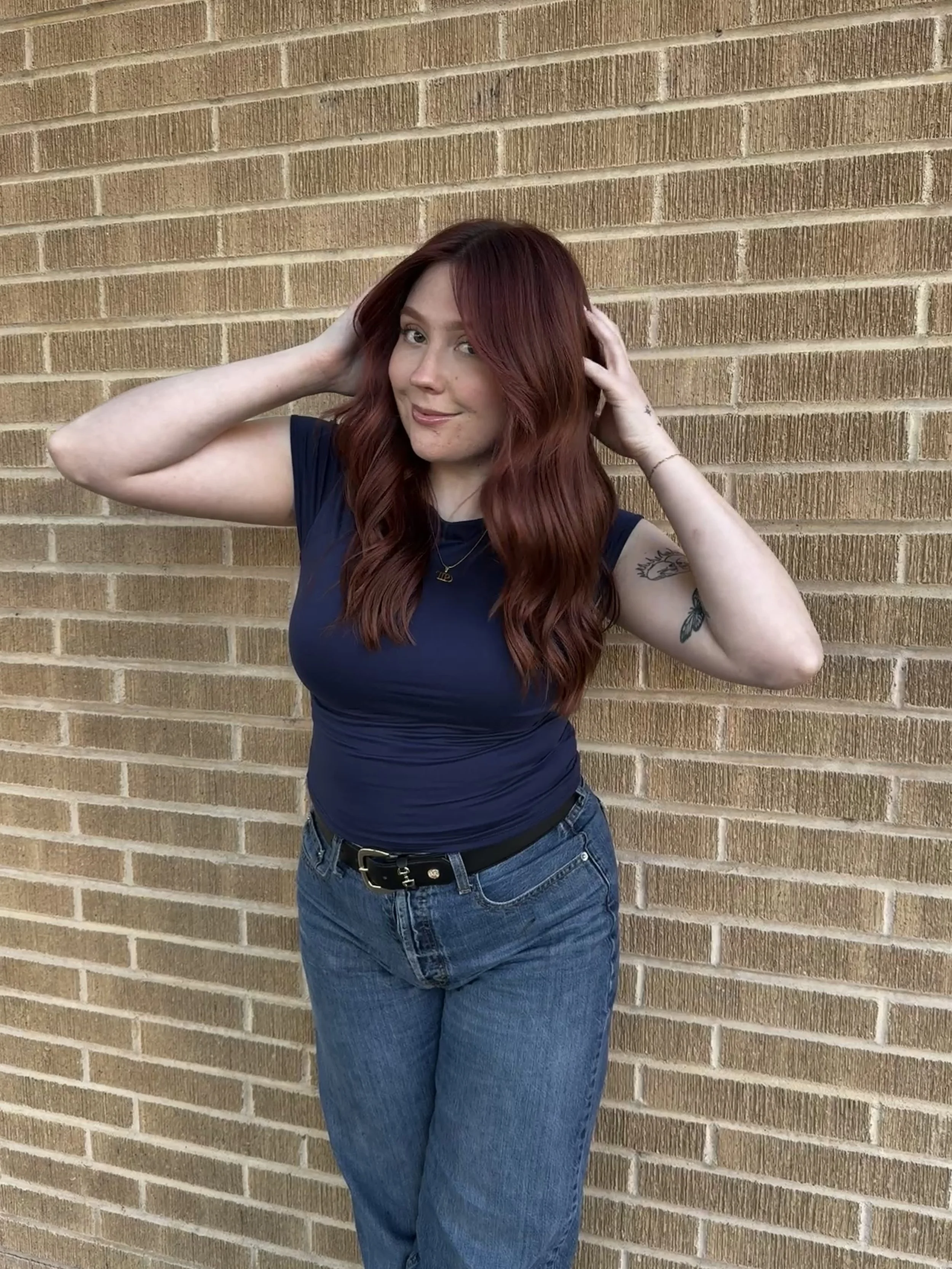 Kassidy Larine, a woman with long red hair in a blue shirt and jeans, standing against a brick wall, smiling, with her hands on her head.