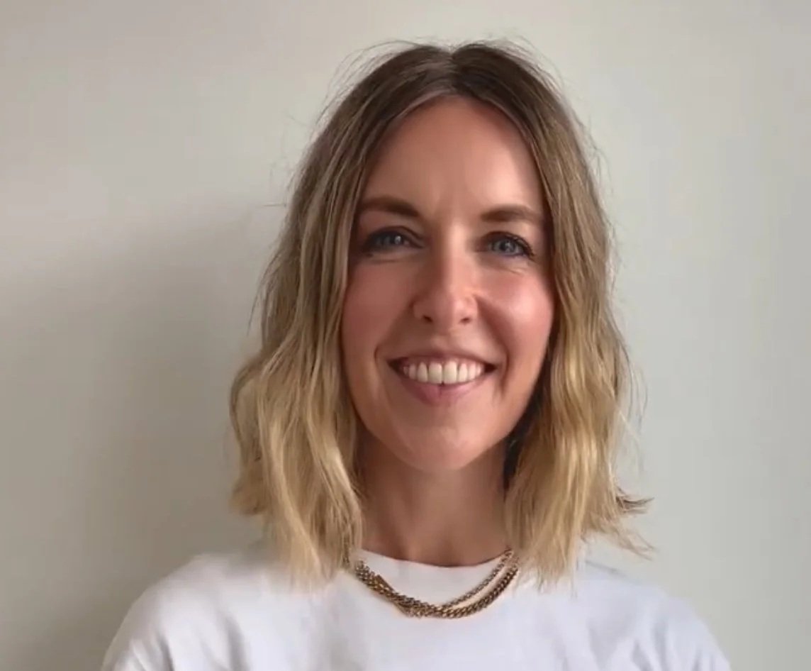 A woman with shoulder-length blonde hair smiling and wearing a white shirt and gold necklace, standing against a plain light-colored wall.