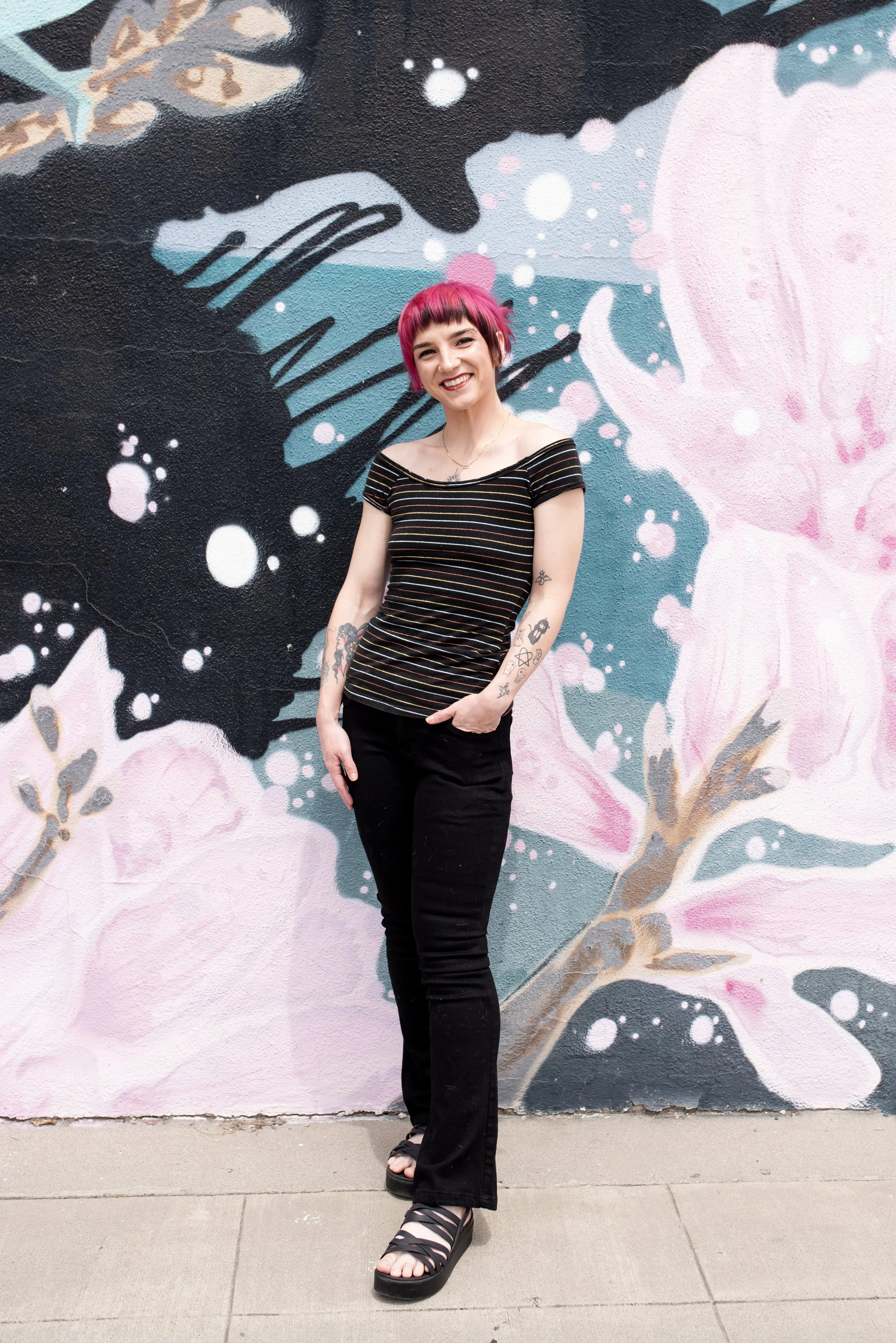 A young woman with pink hair and tattoos on her arms, standing in front of a colorful mural with flowers and abstract patterns, smiling at the camera.