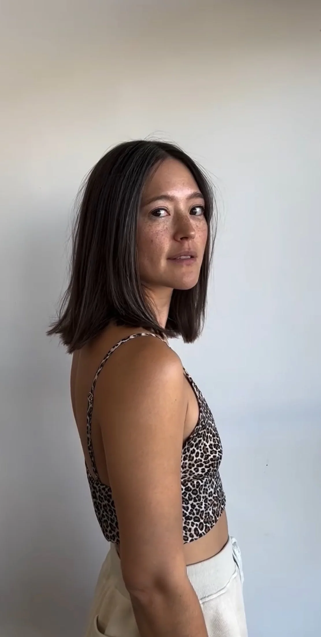 A young woman with shoulder-length dark hair and freckles, wearing a leopard print spaghetti strap top and white high-waisted pants, standing against a plain white wall, looking over her shoulder at the camera.